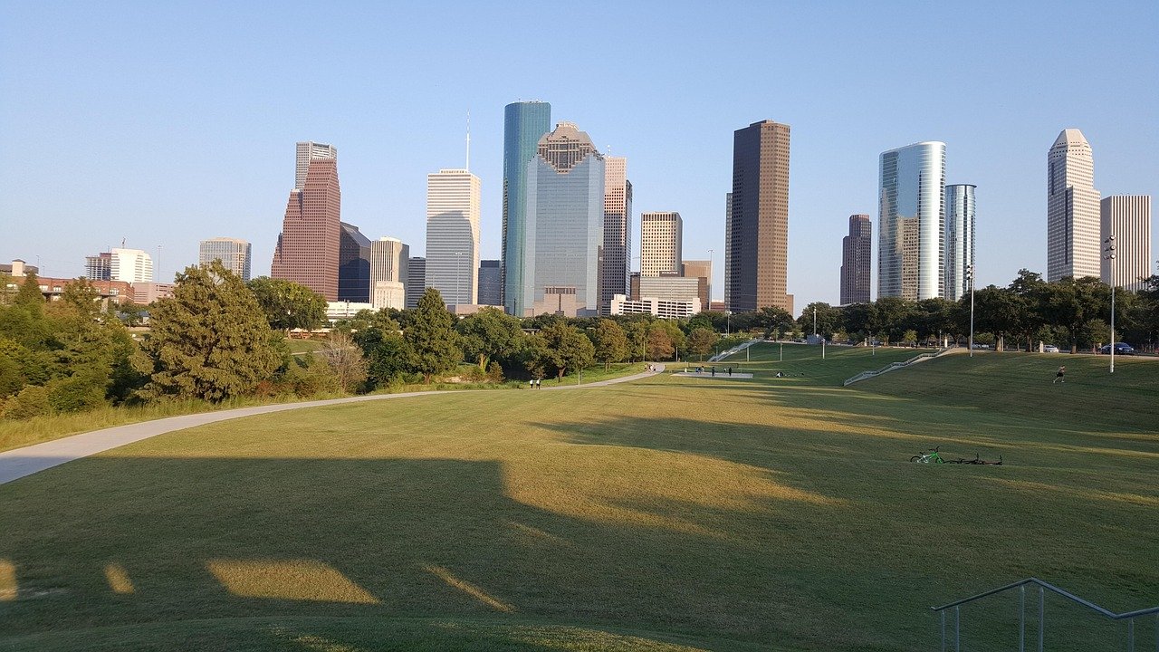 Urban park with green grass, walking path, and city skyline with tall skyscrapers in the background under clear blue sky.