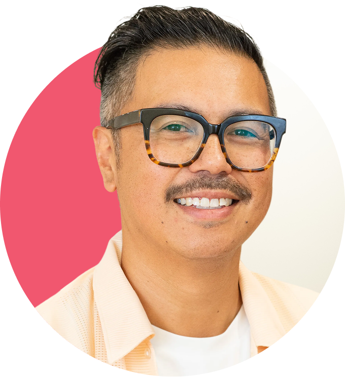 A man with short dark hair, glasses with tortoiseshell frames, and a mustache smiling in front of a background split between pink and white.