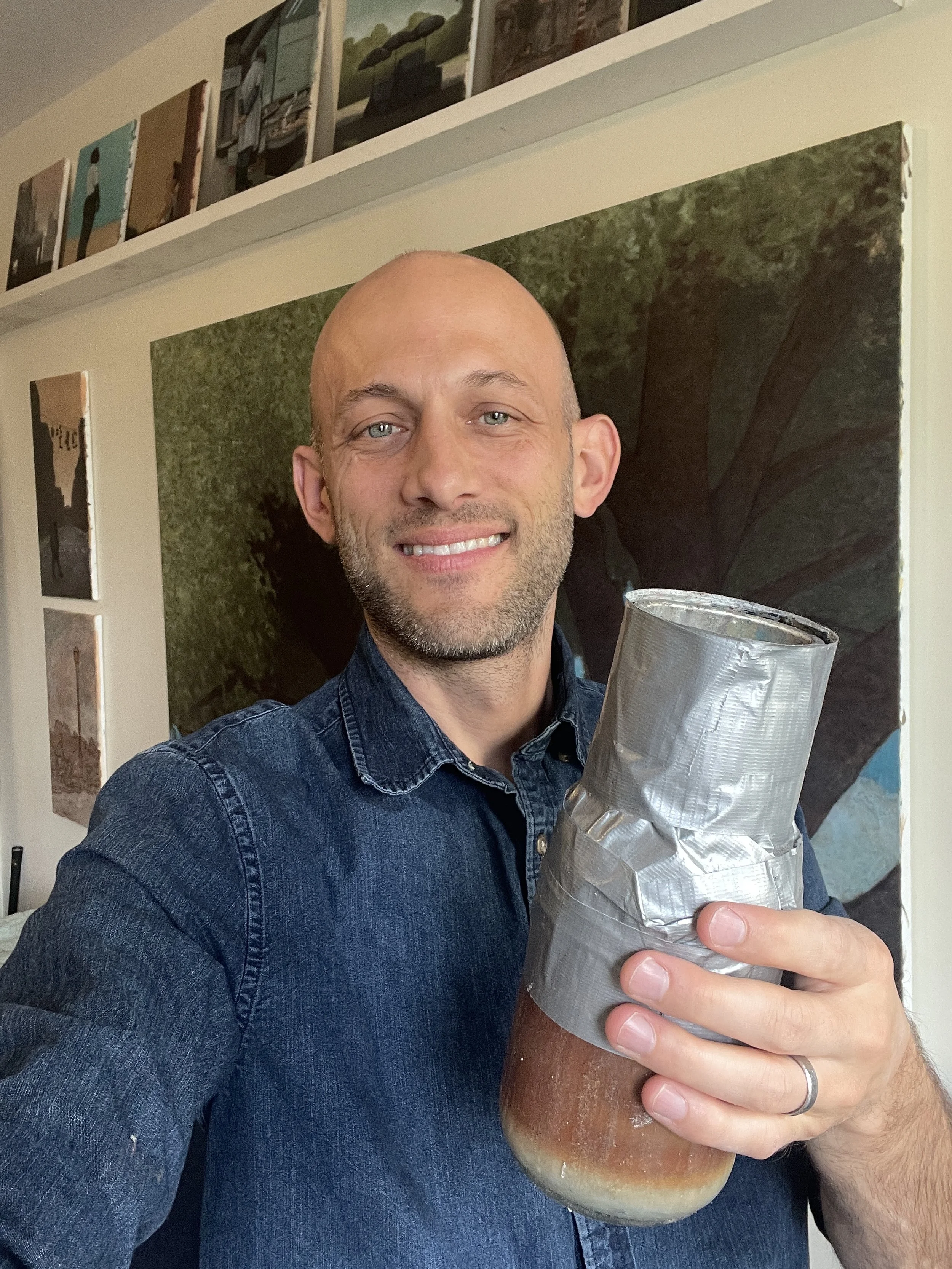 I like what AI wrote here, so I will keep it > A man smiling and holding a jar of homemade beer, wearing a denim shirt, inside a room with artwork on the walls.