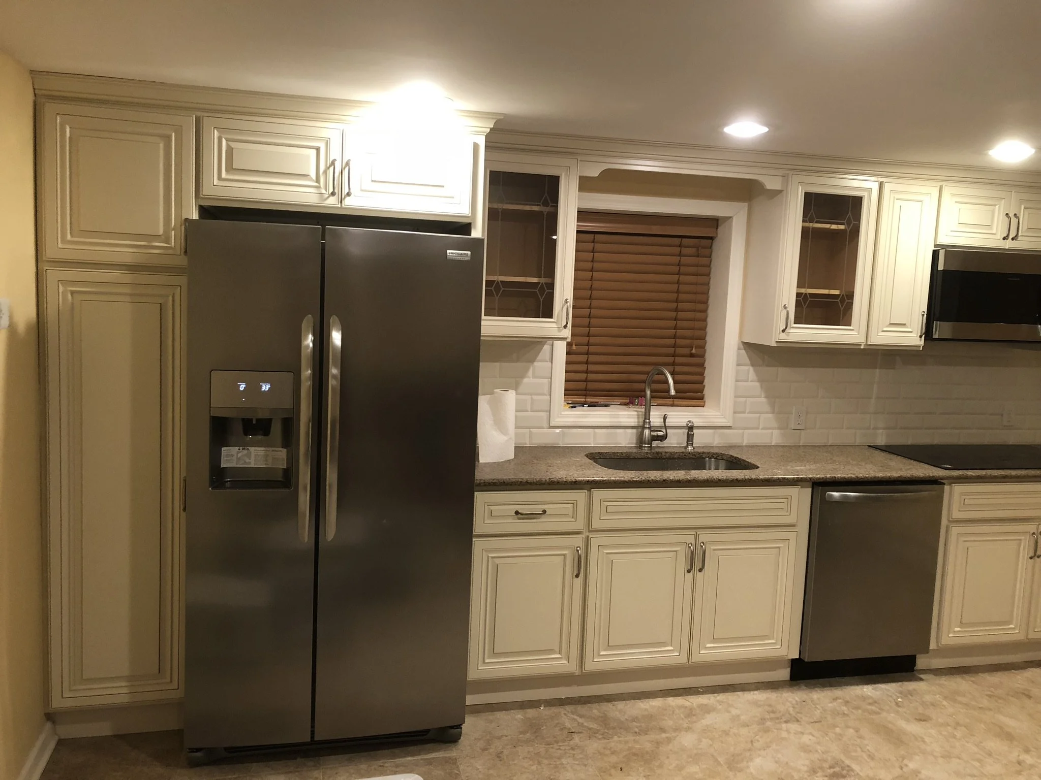 Kitchen with stainless steel refrigerator, white cabinets, granite countertop, sink under window with brown blinds, microwave, and dishwasher.