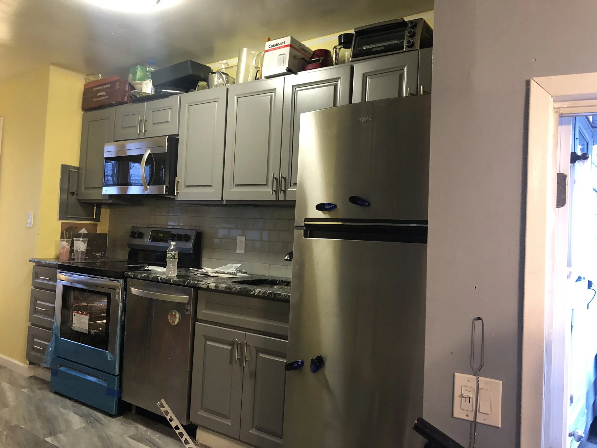 A kitchen with gray cabinets, stainless steel appliances, a marble countertop, and various items on top of the cabinets, including boxes, a kettle, and a toaster. There is a door on the right side.