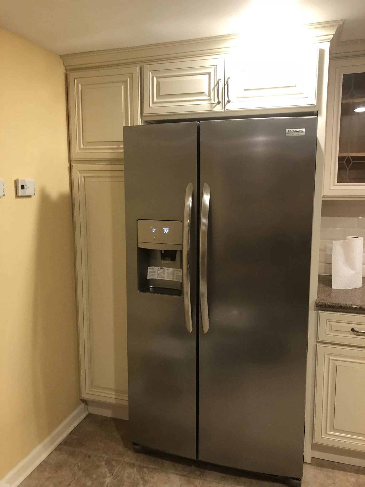 Stainless steel Frigidaire side-by-side refrigerator in a kitchen with cream cabinetry and yellow wall.