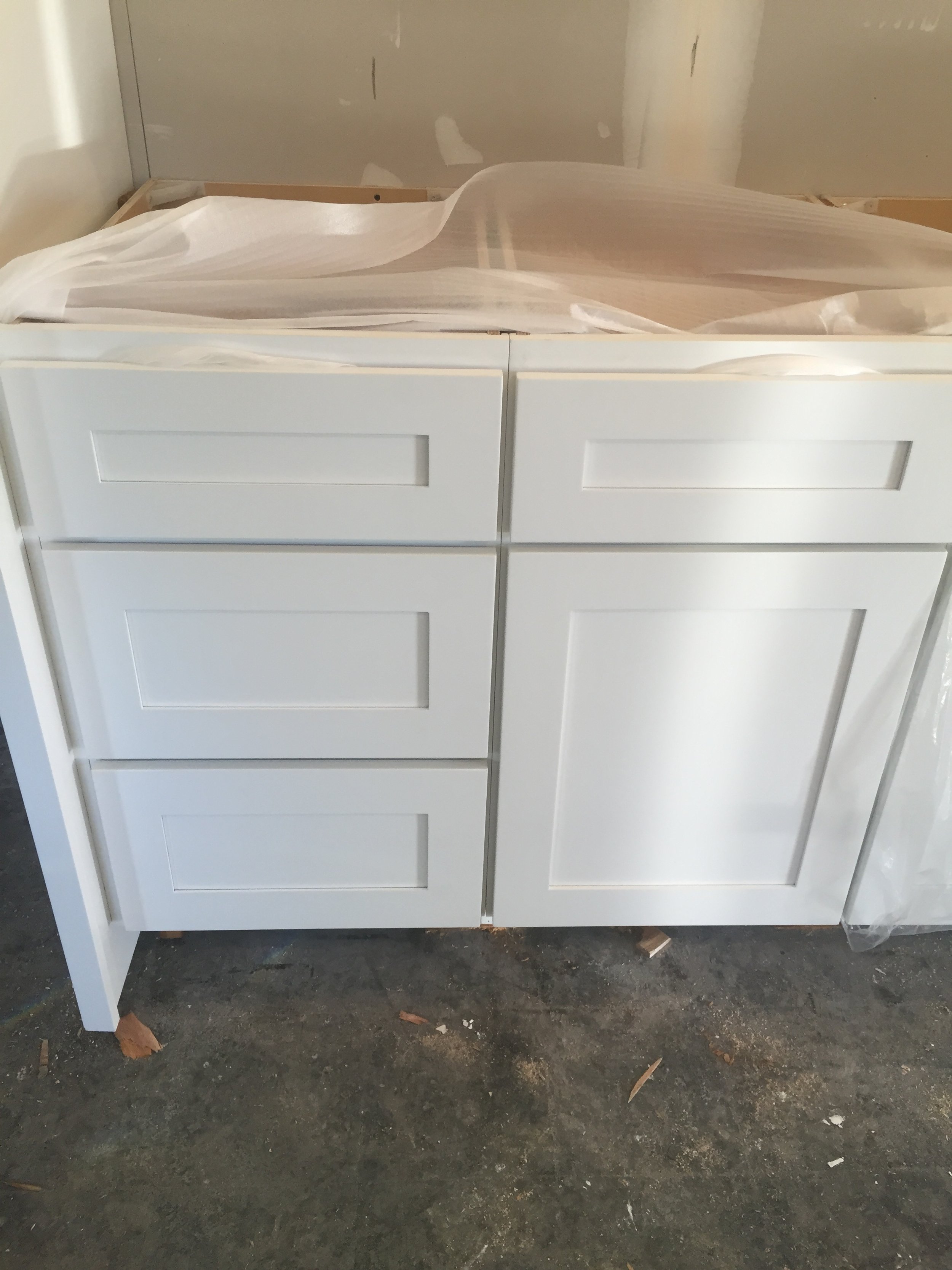 White kitchen cabinet with four drawers on the left and a large door on the right, placed on a concrete floor, partially covered in plastic and paper.
