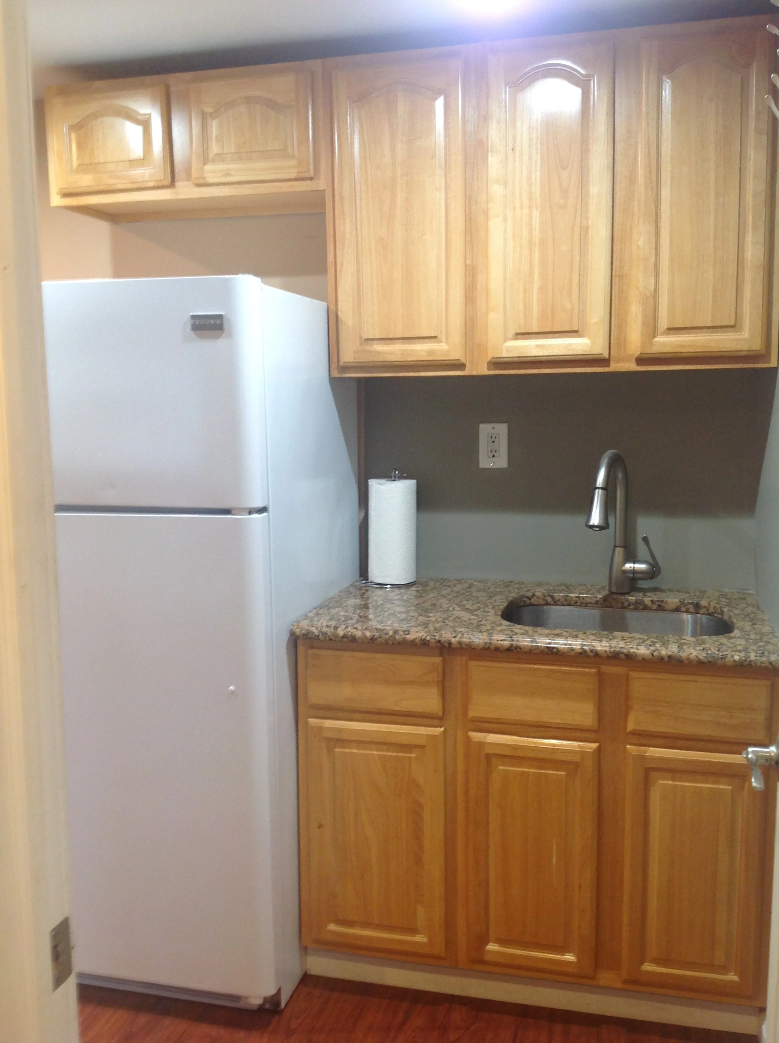 A small kitchen with wooden cabinets, a granite countertop, and a white refrigerator. There is a sink with a modern faucet and a roll of paper towels on the counter.