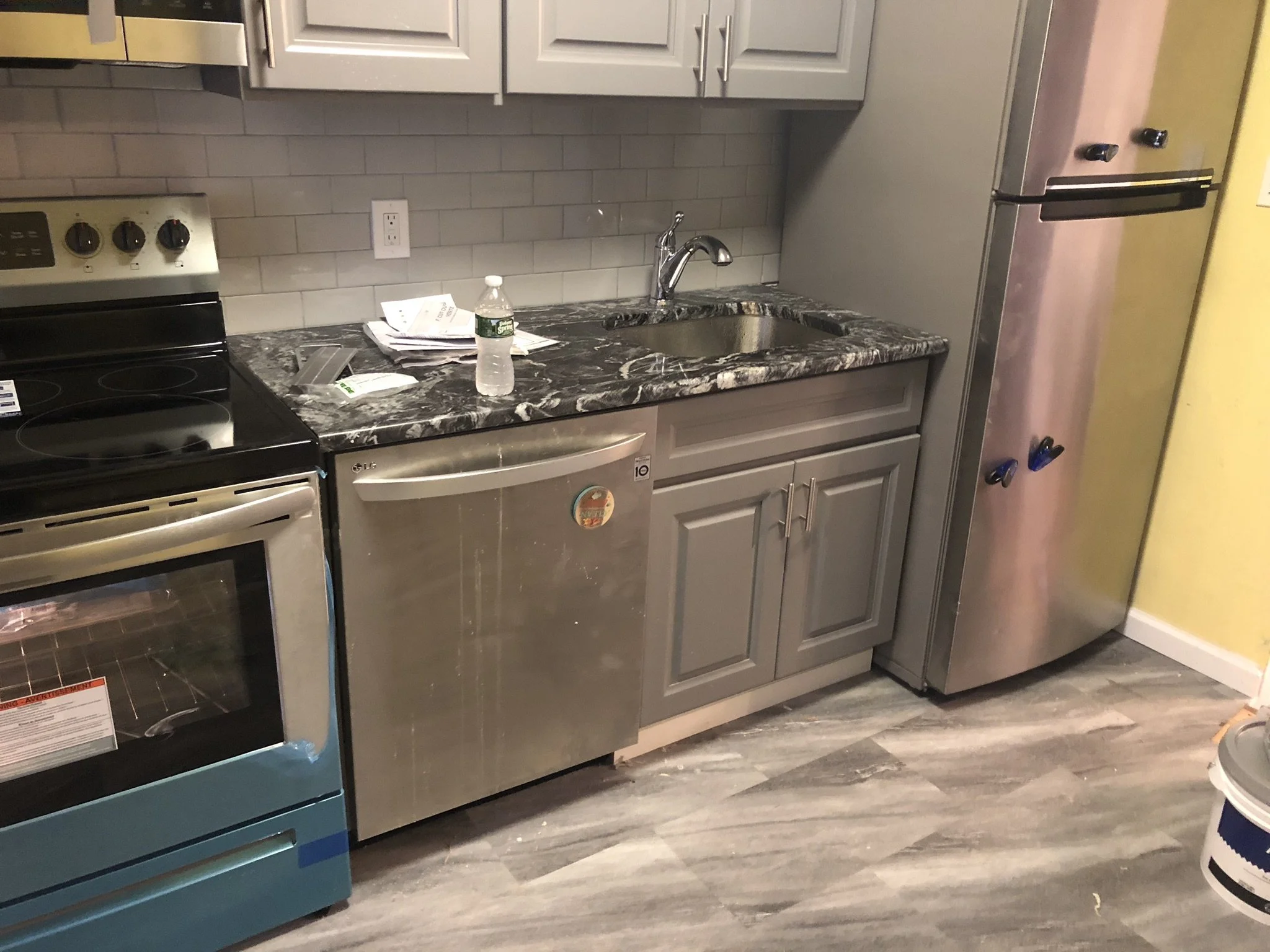 Kitchen with a black stove, a stainless steel dishwasher, gray cabinets, a marble countertop, a sink, a water bottle, some paper, and a stainless steel refrigerator.