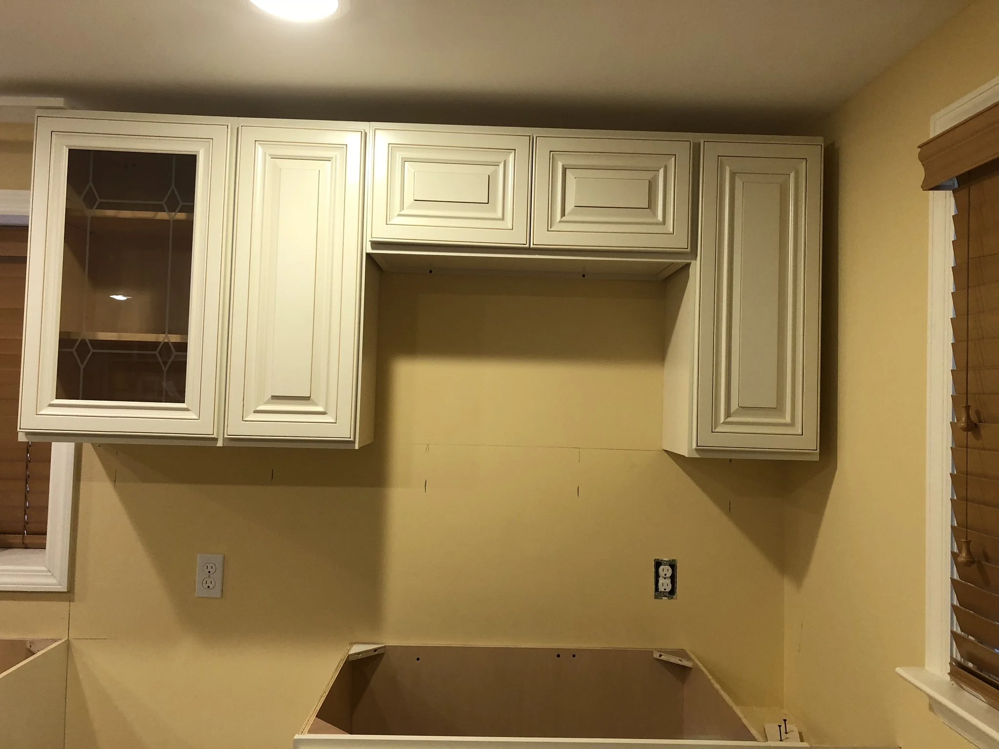 Kitchen with cream-colored cabinets, some with glass doors, and yellow walls, showing an unfinished space for a stove or oven.