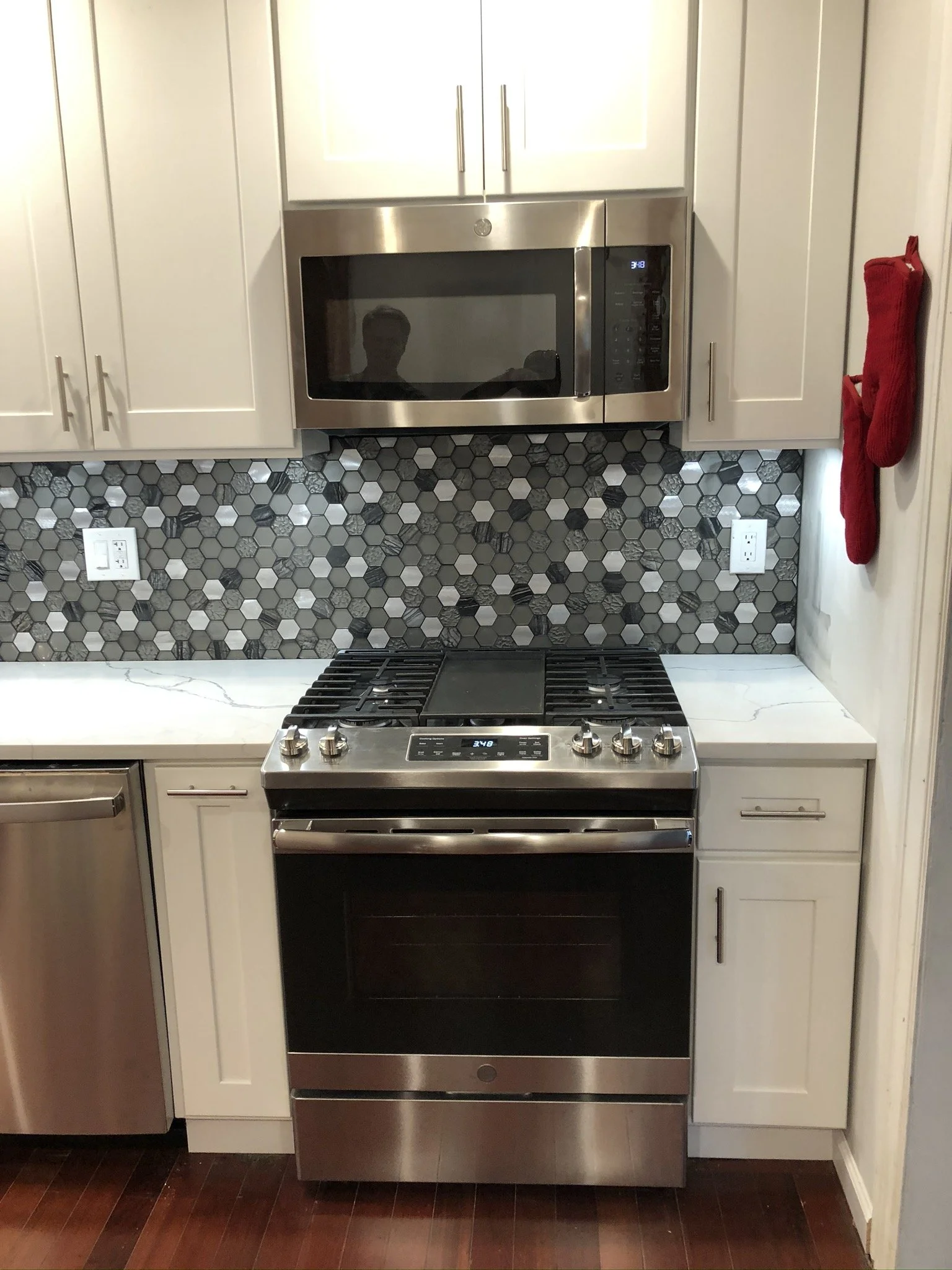 Kitchen with white cabinets, hexagon tile backsplash in gray tones, a stainless steel microwave, and a stainless steel stove with oven. There is a red oven mitt hanging on the side of the cabinet.