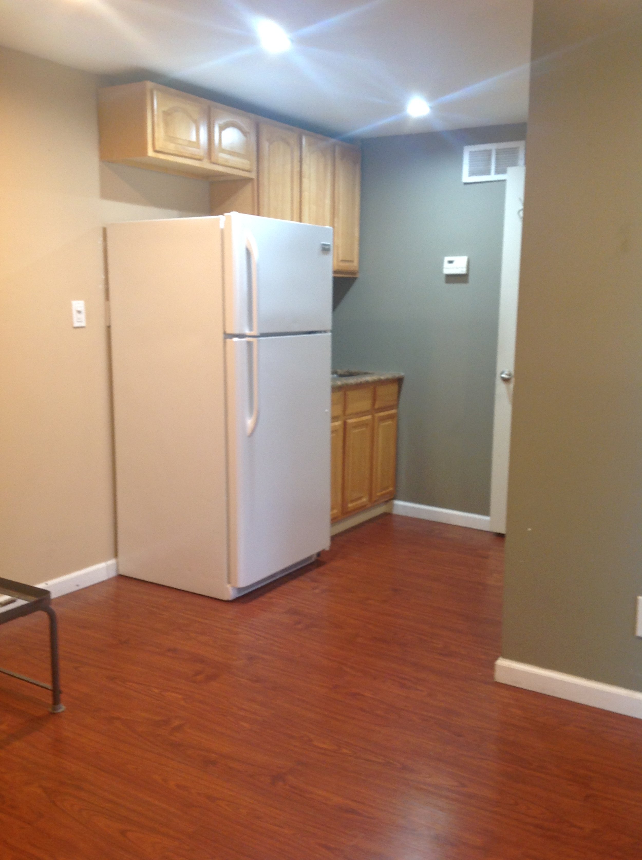 Image of a kitchen with a white refrigerator, wooden cabinets, and hardwood flooring. The walls are painted in neutral colors, and ceiling lights are on.
