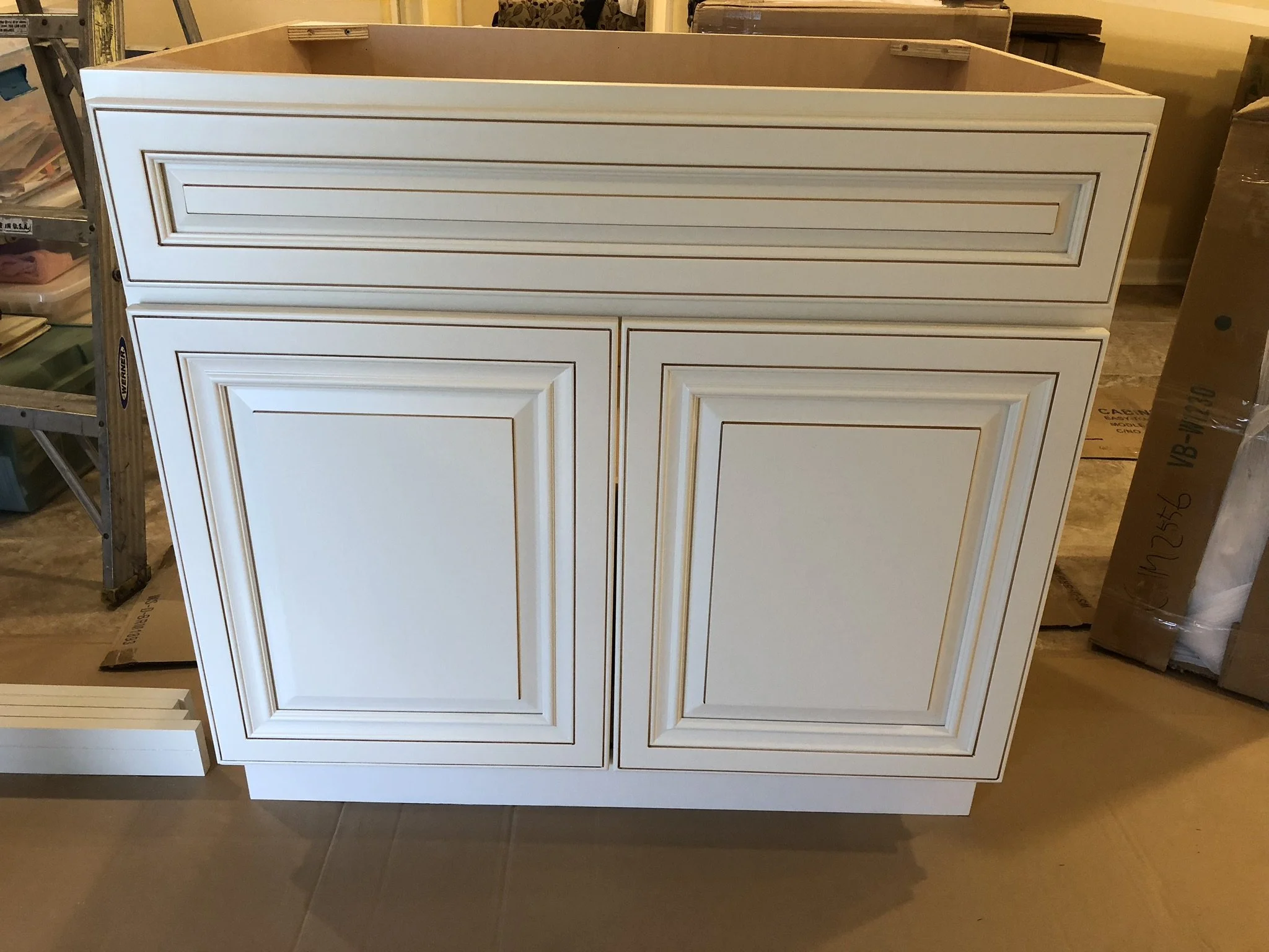 A white cabinet with decorative molding on the doors and drawer, situated in a workshop or storage area.