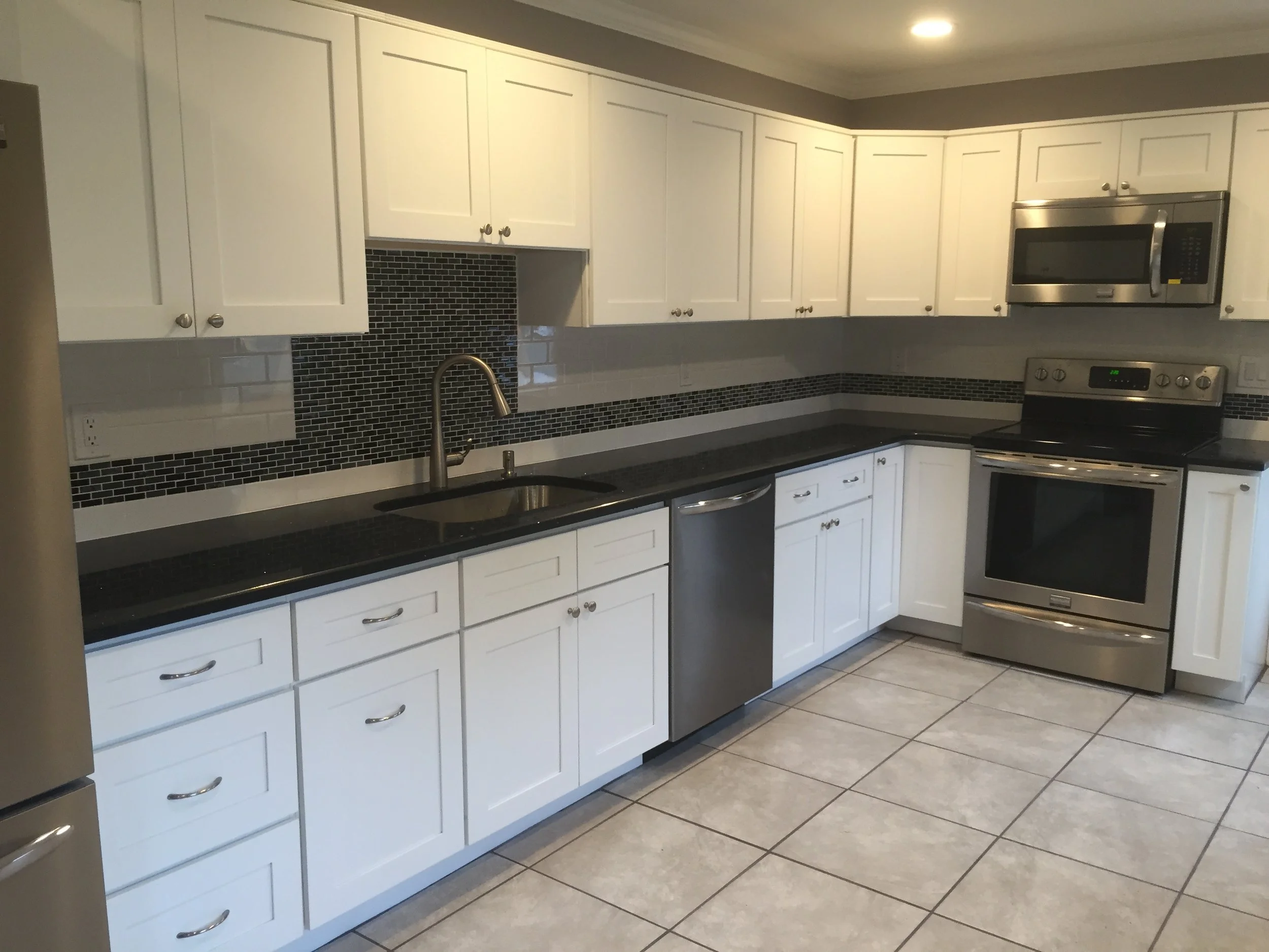 Modern kitchen with white cabinets, black countertop, stainless steel appliances including microwave, stove, and dishwasher, and a black tile backsplash.