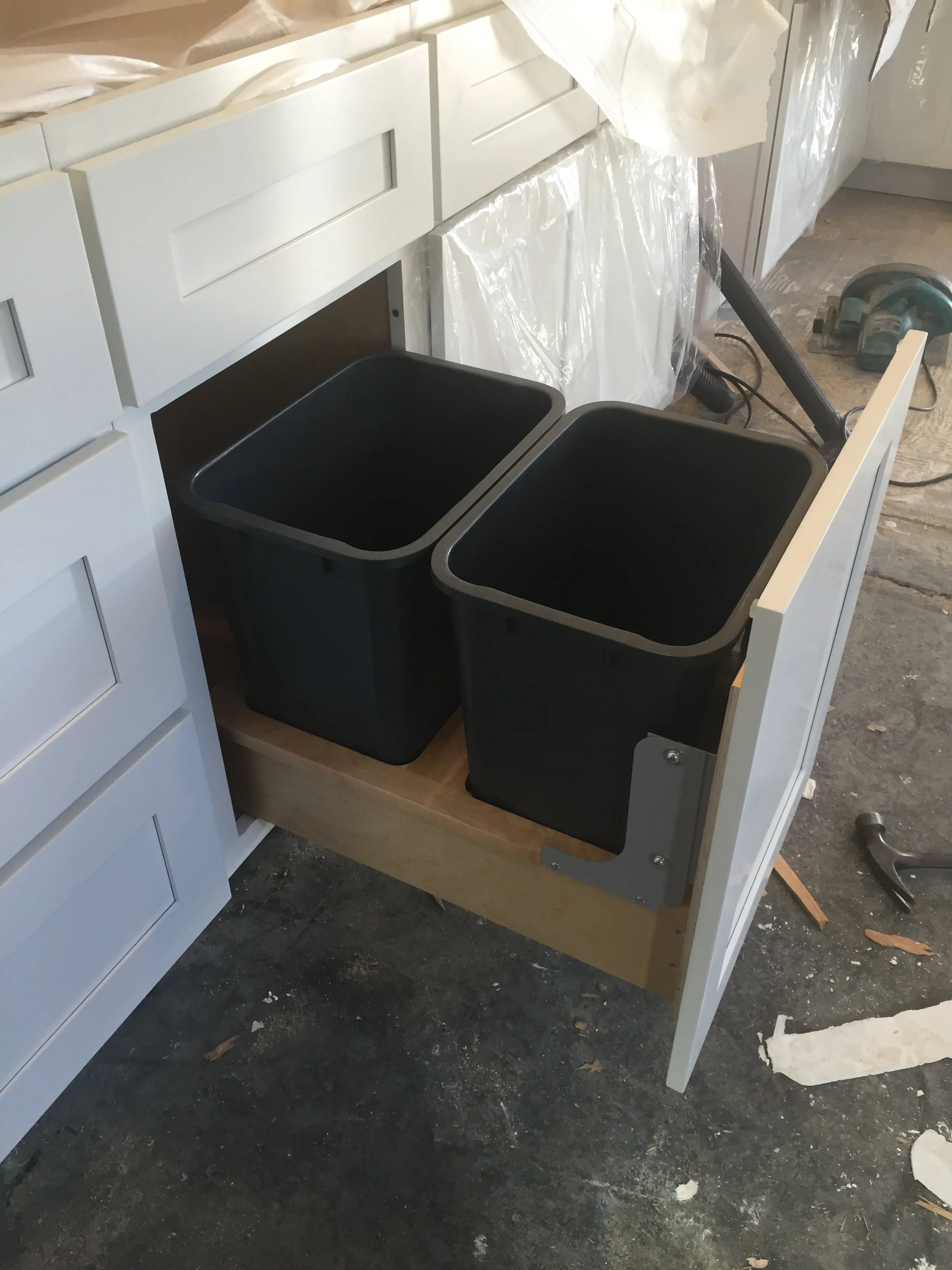 Under-construction kitchen drawer with two black trash bins inside, white cabinets above and a power drill on the floor nearby.