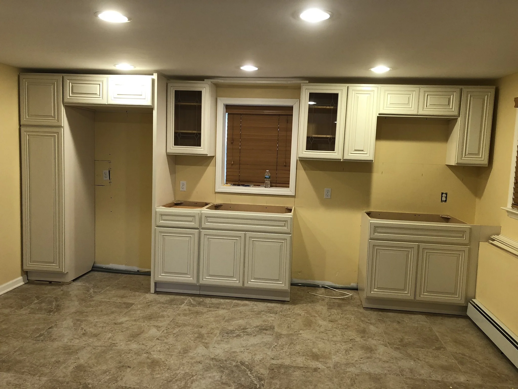 Partially installed kitchen cabinets with no appliances, yellow walls, a window with brown blinds, and unfinished countertops, with electrical outlets and cables visible.