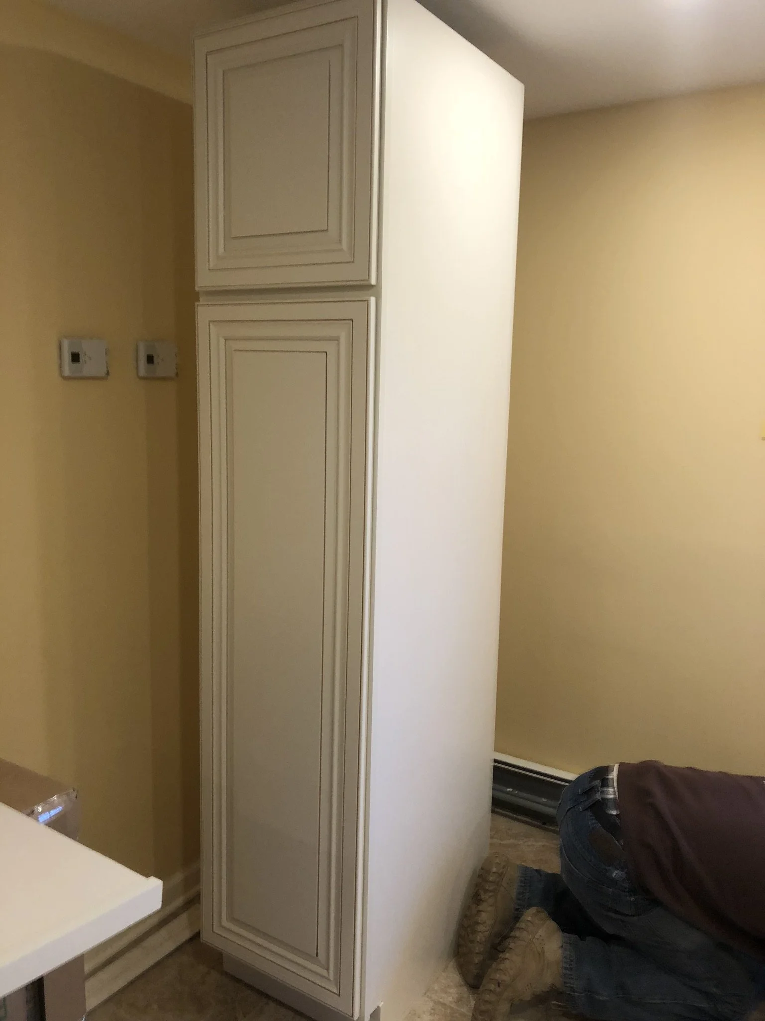 Person kneeling on the floor installing or repairing a tall kitchen cabinet in a room with yellow walls, baseboard heater, and thermostat controls on the wall.