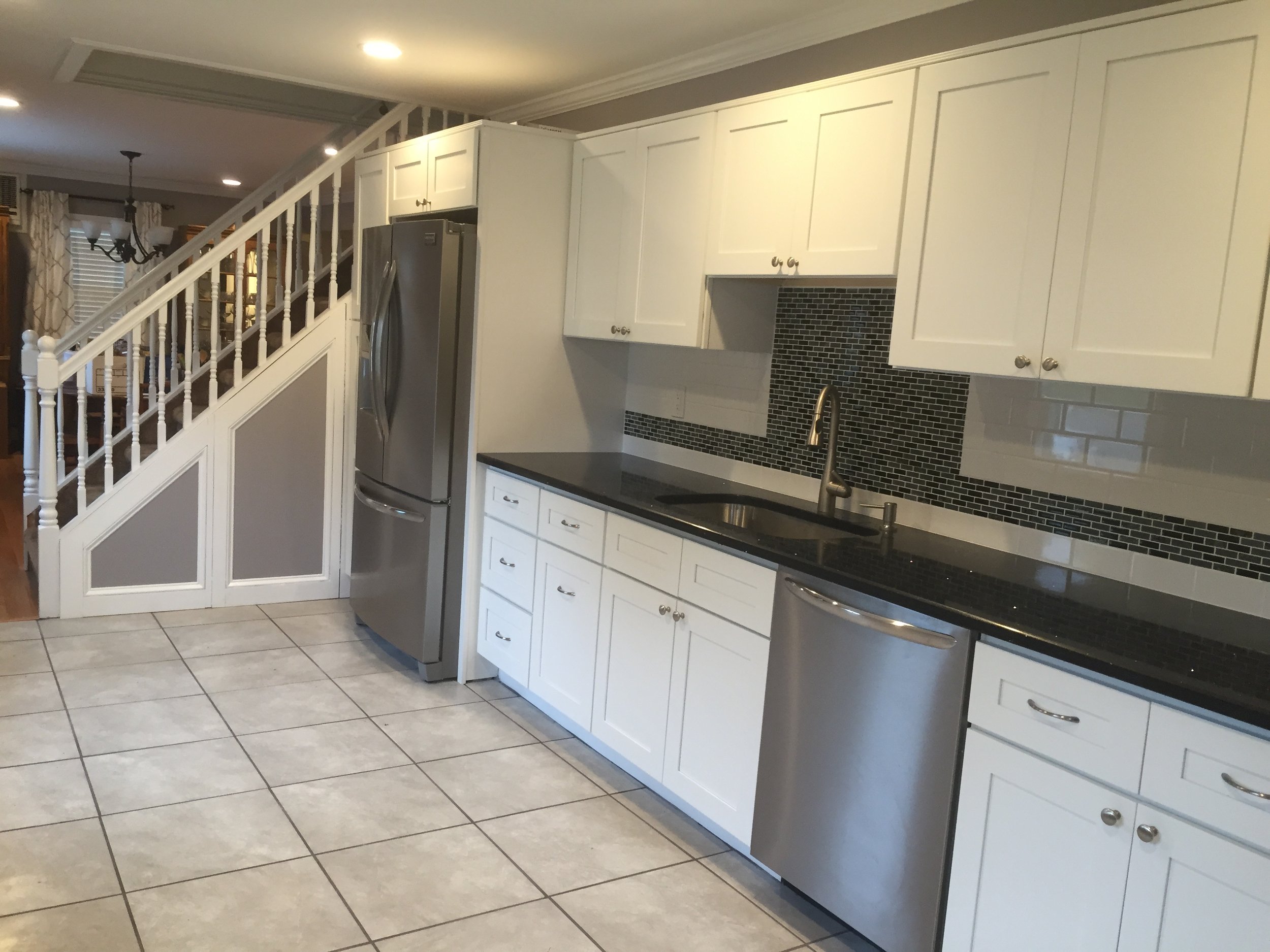 Modern kitchen with white cabinets, black countertop, stainless steel refrigerator and dishwasher, black tile backsplash, and tiled floor.