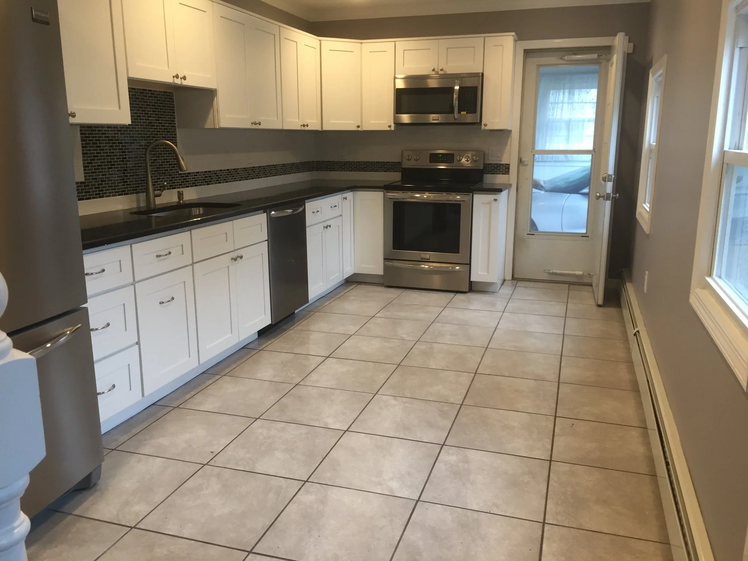 Empty kitchen with white cabinets, black countertops, and beige tiled floor. Stainless steel appliances including a refrigerator, microwave, oven, and dishwasher. There is a window and a door with a window leading outside.