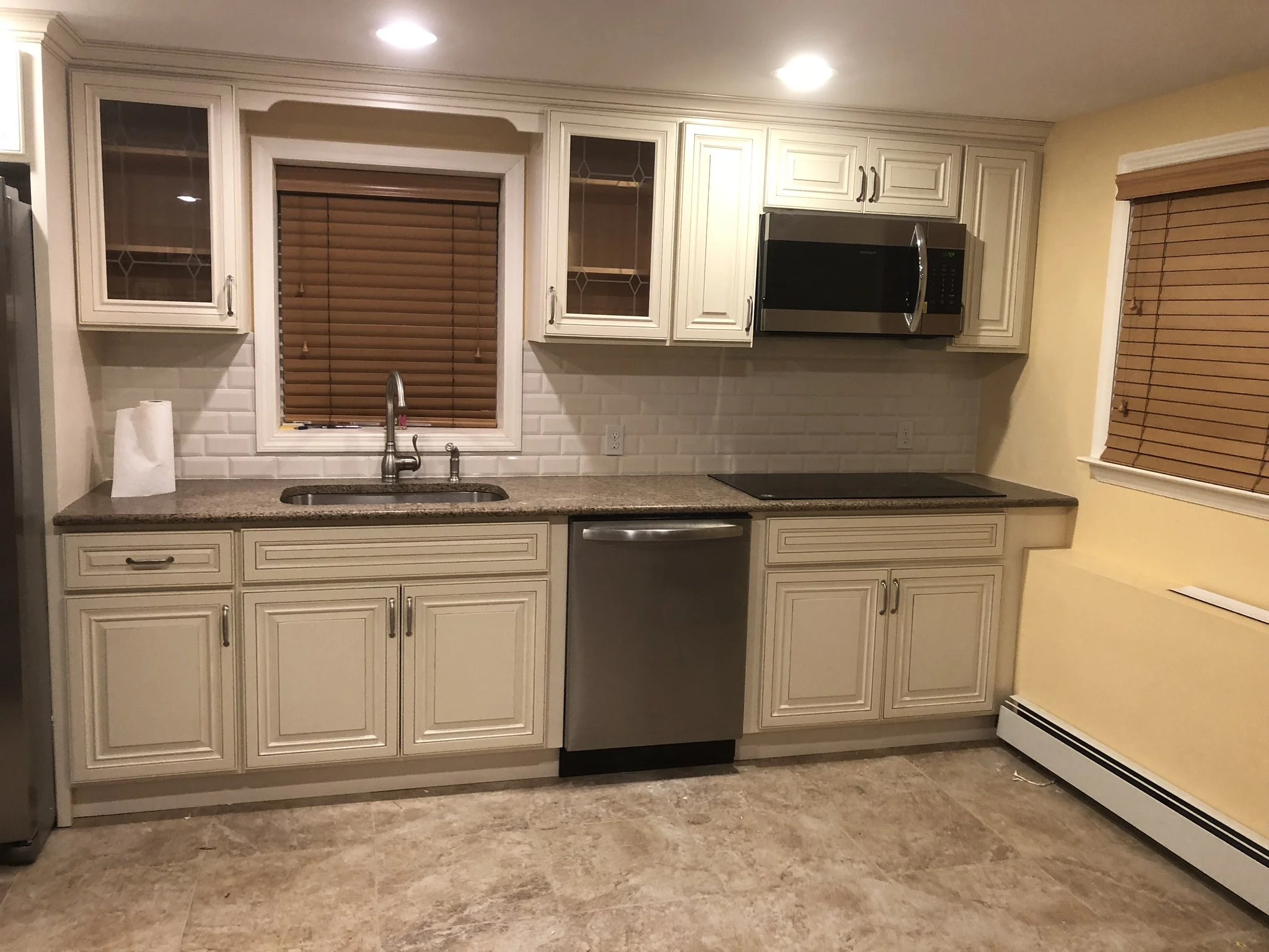 Kitchen with white cabinets, granite countertops, a window with brown blinds, a stainless steel dishwasher, a stainless steel microwave, and a beige wall with a window and a radiator.