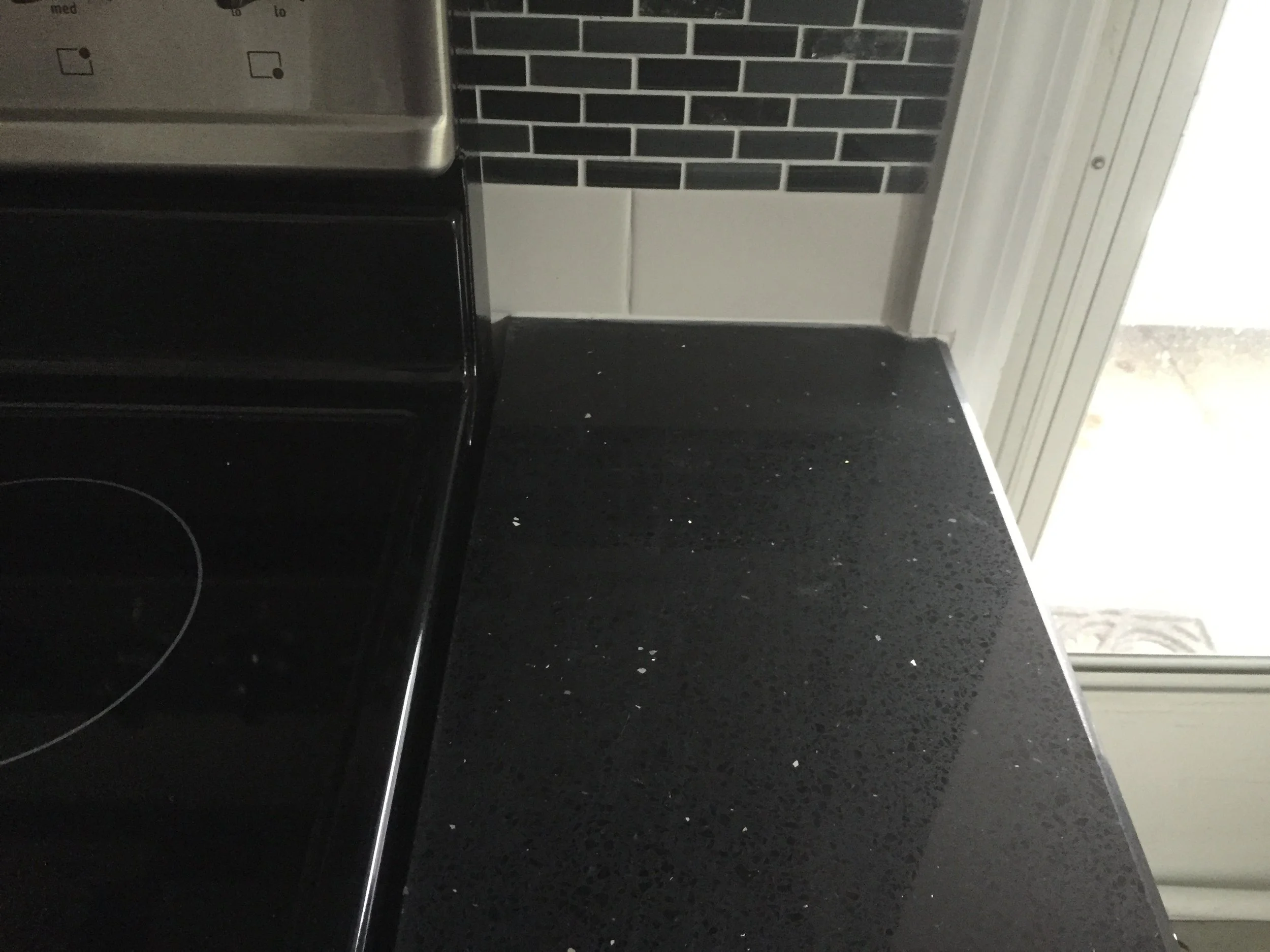 Close-up of a black kitchen countertop with some white specks, next to a black electric stove and a window with white framing, showing a glimpse of an outdoor area.