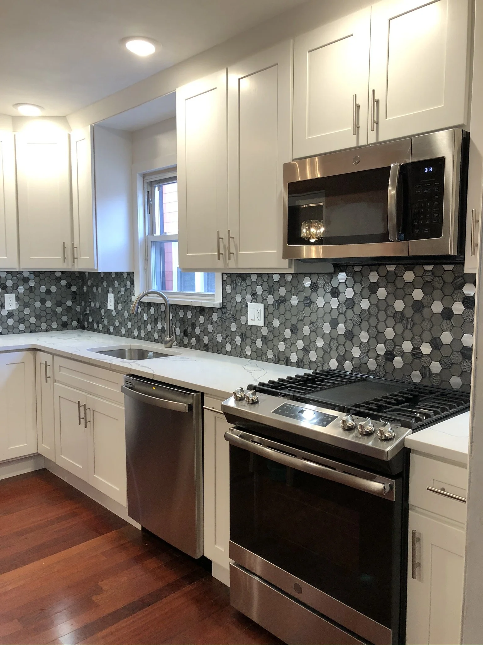 Modern kitchen with white cabinets, black hexagonal tile backsplash, stainless steel appliances including microwave, oven, and dishwasher, window above sink, wood floor, and ceiling lights.