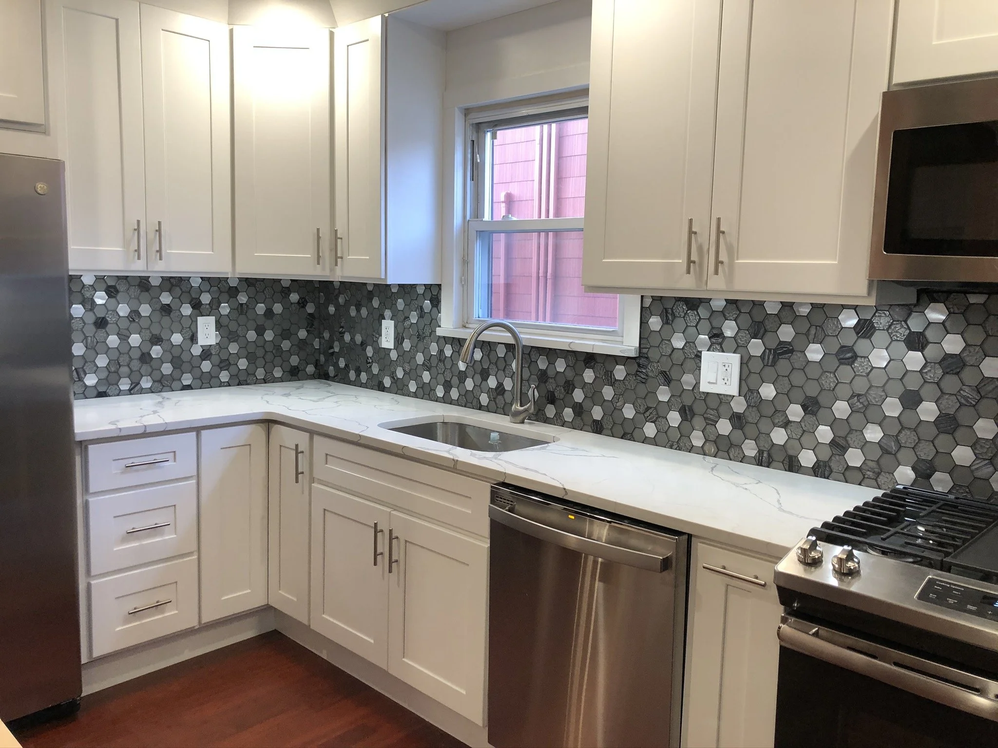 Kitchen with white cabinets, a black and gray hexagonal tiled backsplash, white marble counter, stainless steel appliances, a window above the sink, and hardwood flooring.