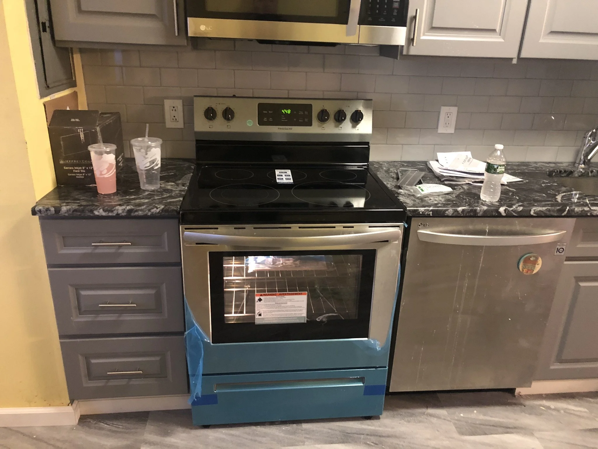 Kitchen with a stainless steel oven, grey cabinets, a black marble countertop, a water bottle, some papers, and drinks in cups on the counter.