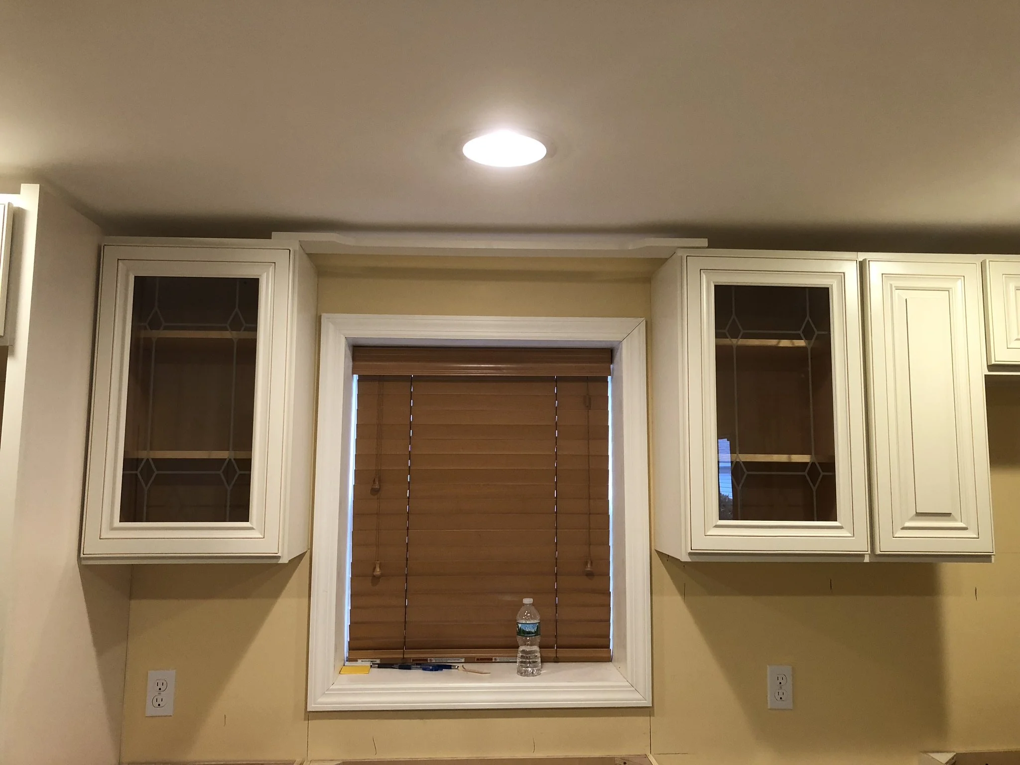 Kitchen wall with a window covered by brown horizontal blinds, white cabinets with glass and solid doors, and two electrical outlets.