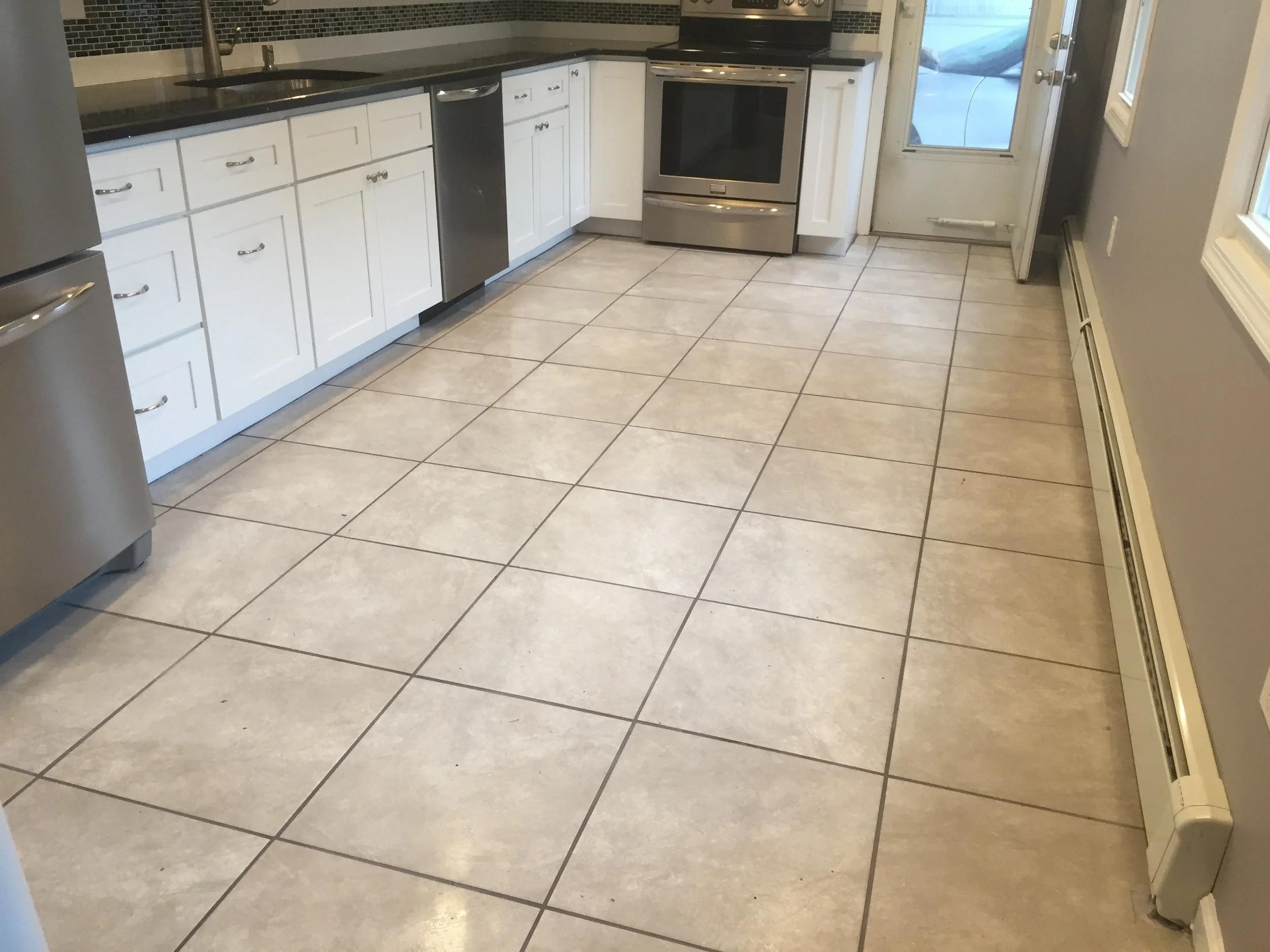 Kitchen with white cabinets, black countertops, stainless steel appliances, beige tiled floor, and a door leading outside.