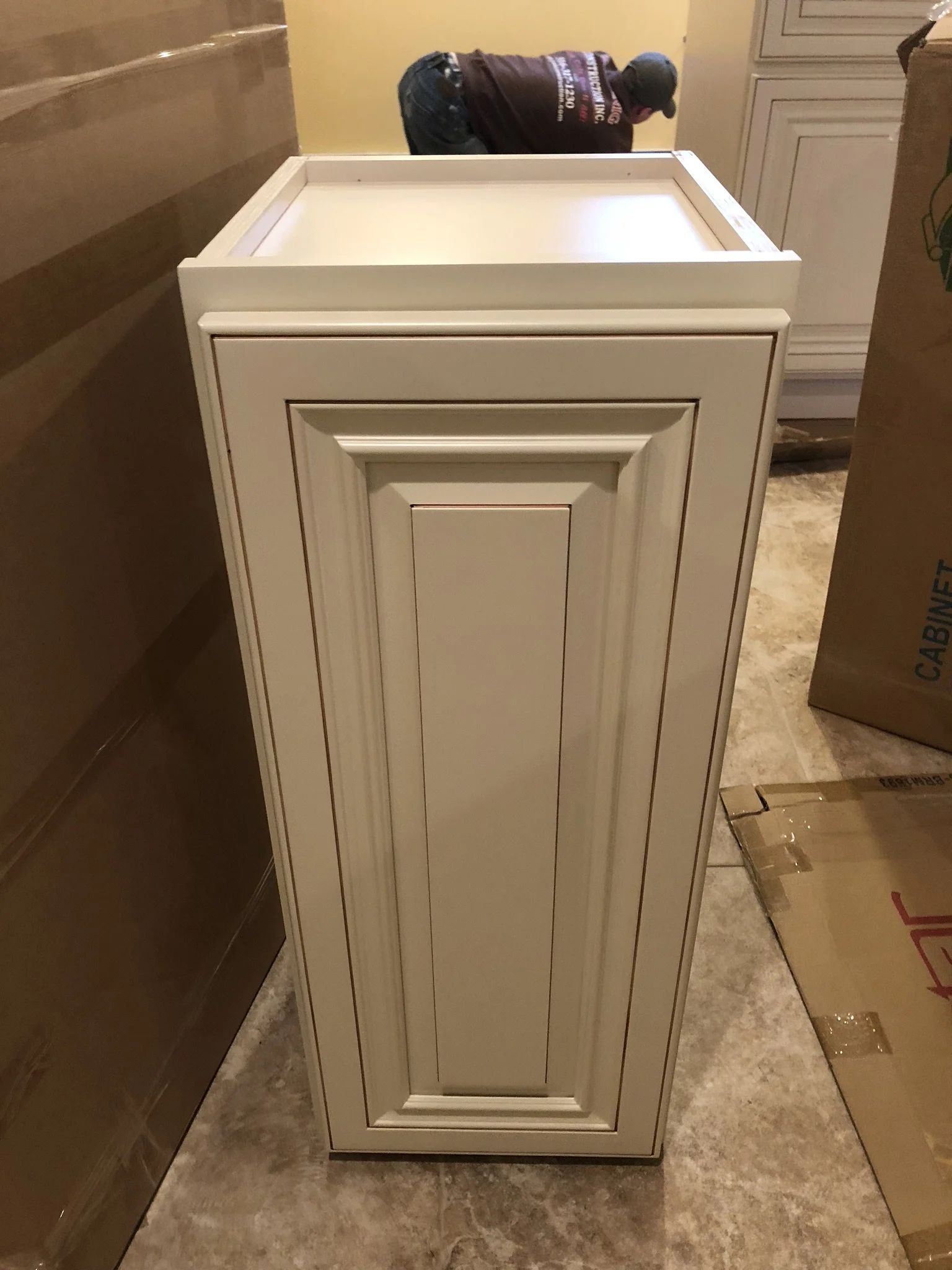 A person in a dark shirt and baseball cap bent over working near a white cabinet or piece of furniture in a kitchen or workshop.