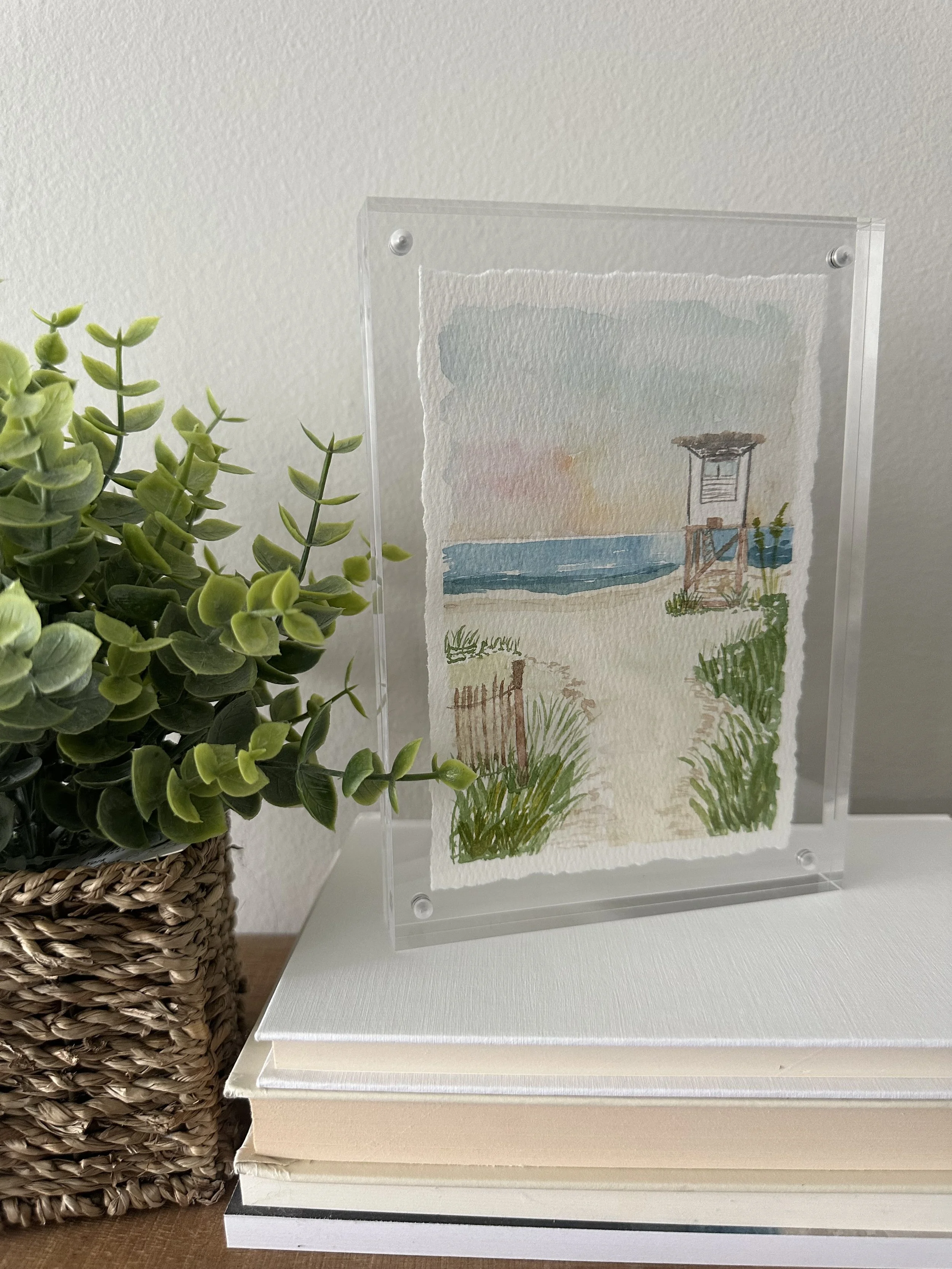 A watercolor painting of a beach scene with a lifeguard tower, sand, grass, and the ocean in the background, displayed on a clear stand on top of a stack of books next to a basket with green plant leaves.