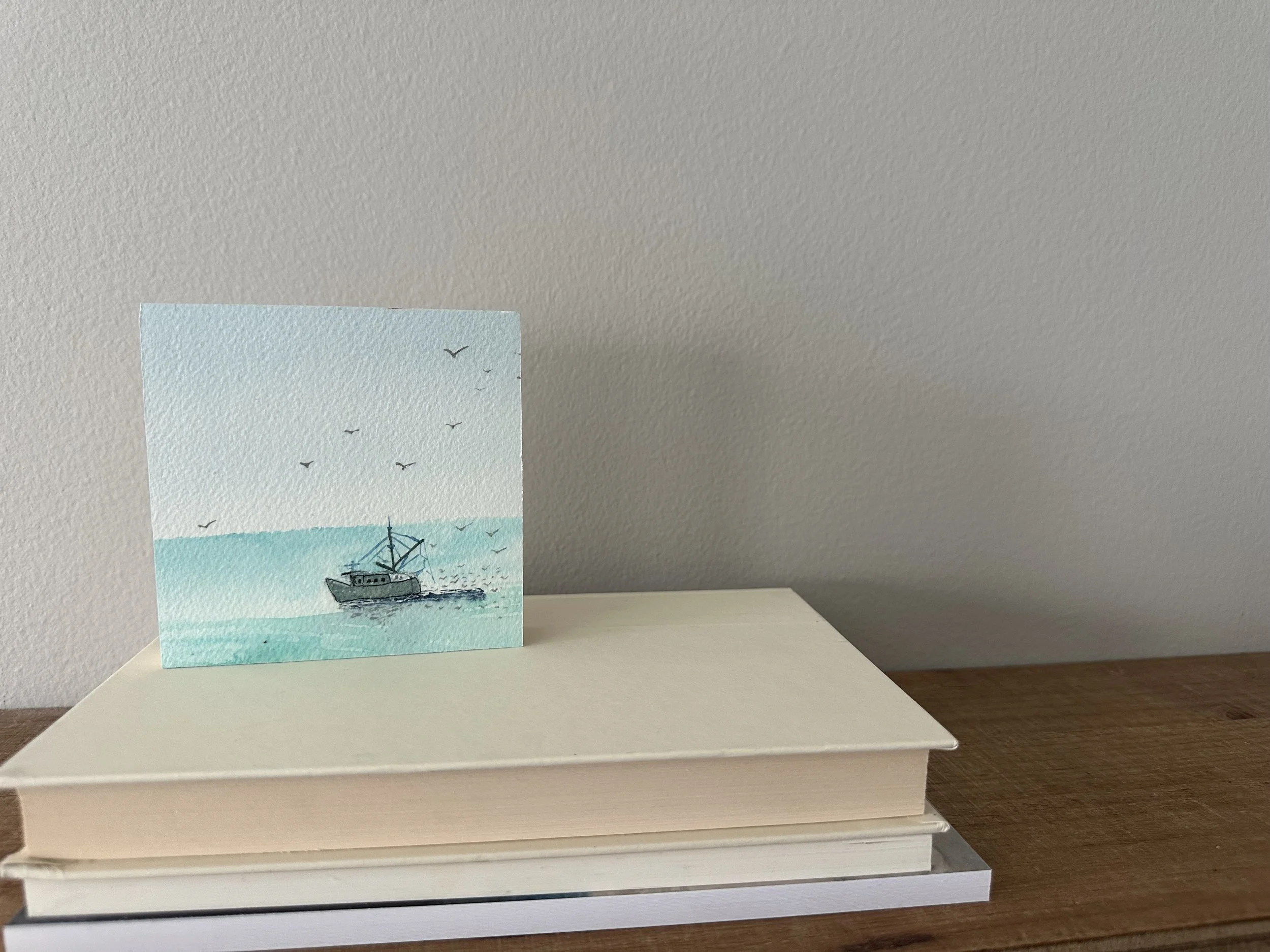 Watercolor painting of a boat on the ocean with several seagulls flying in the sky, placed on top of a stack of two books on a wooden surface against a light-colored wall.