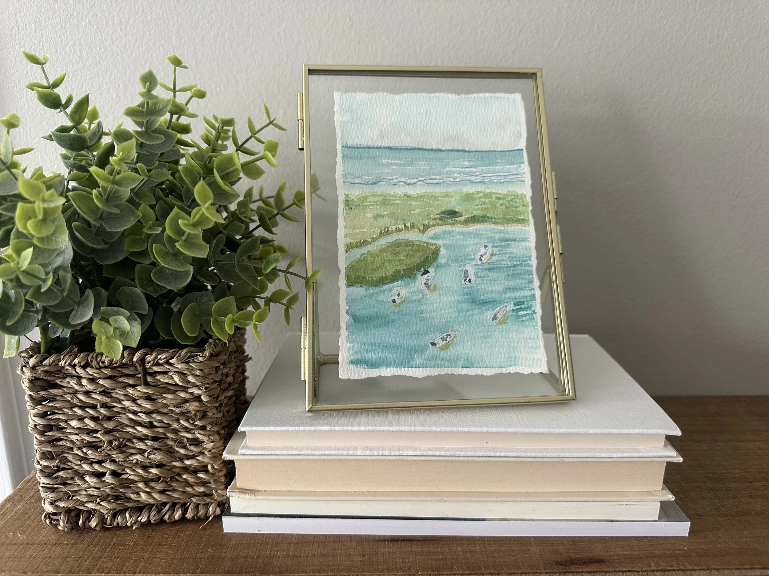 A framed watercolor painting of a coastal scene with water, greenery, and seagulls, sitting on a white bookstacks and next to a green leafy plant in a woven basket on a wooden surface.