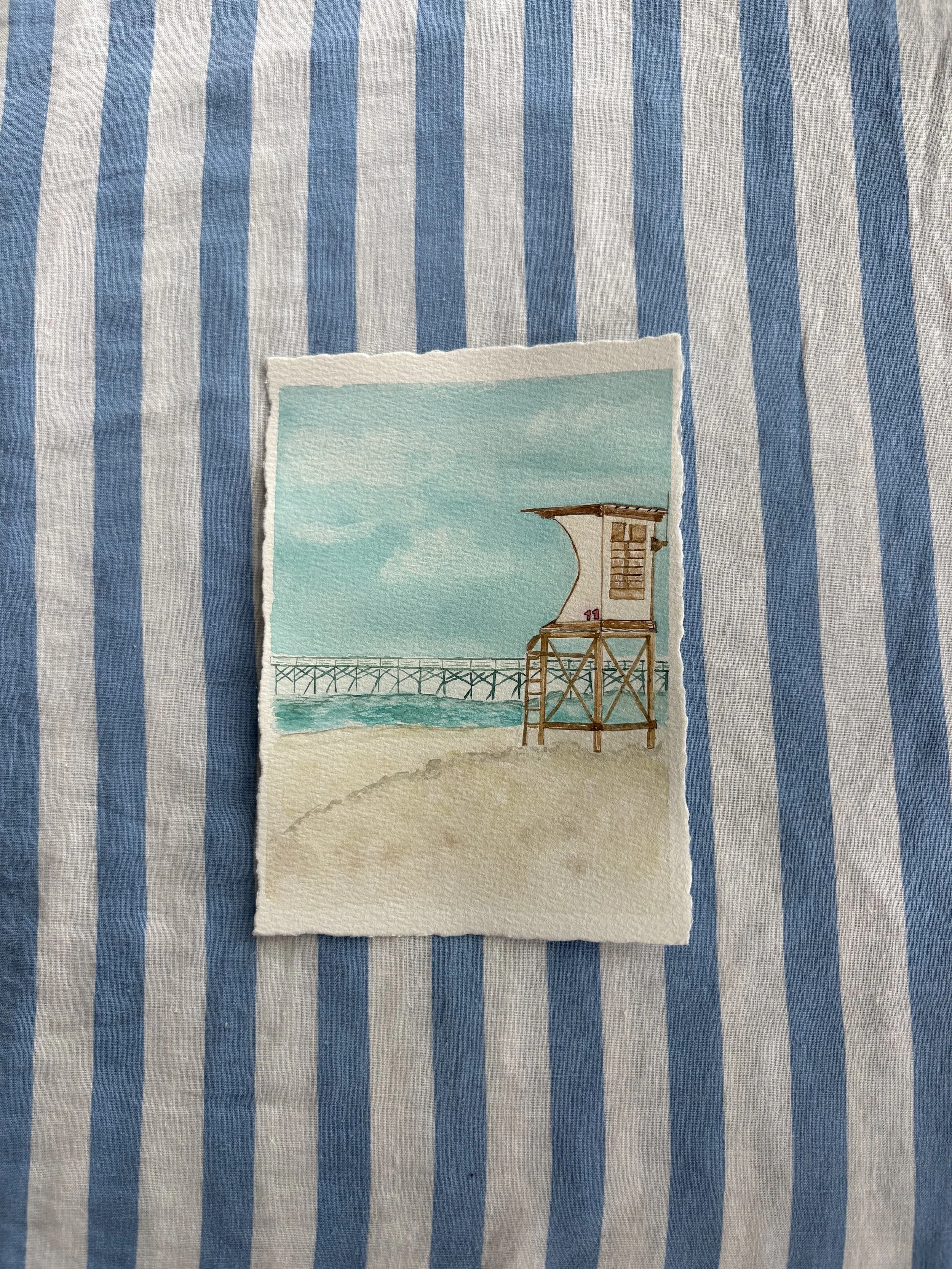 Watercolor painting of a lifeguard tower on a sandy beach with a pier in the background and a cloudy sky.