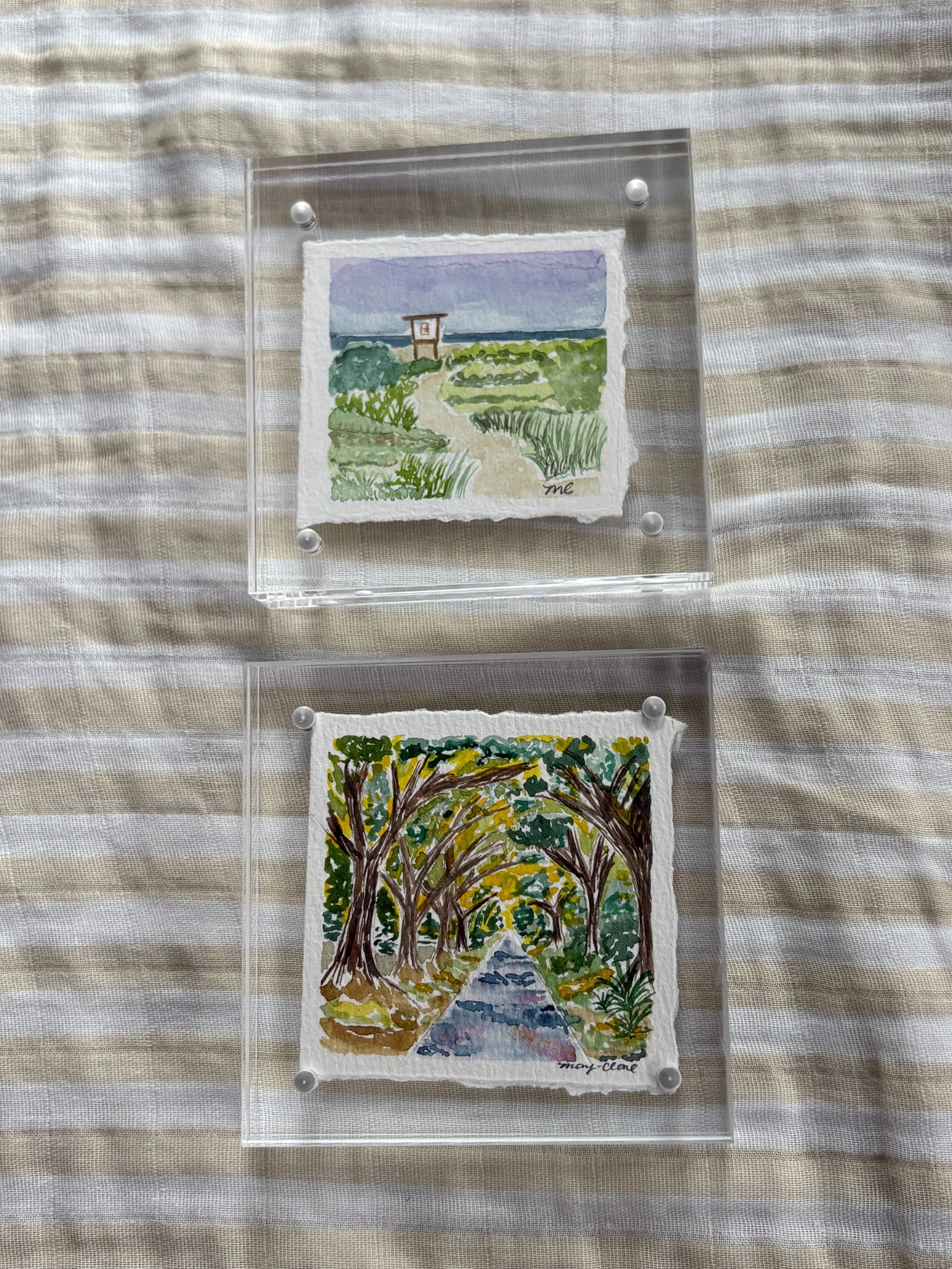 Two small watercolor paintings framed with clear plastic mounts, featuring nature scenes. The top painting depicts a landscape with a pathway, greenery, and a blue sky, signed 'me'. The bottom painting shows a tree-lined path in autumn colors, with g