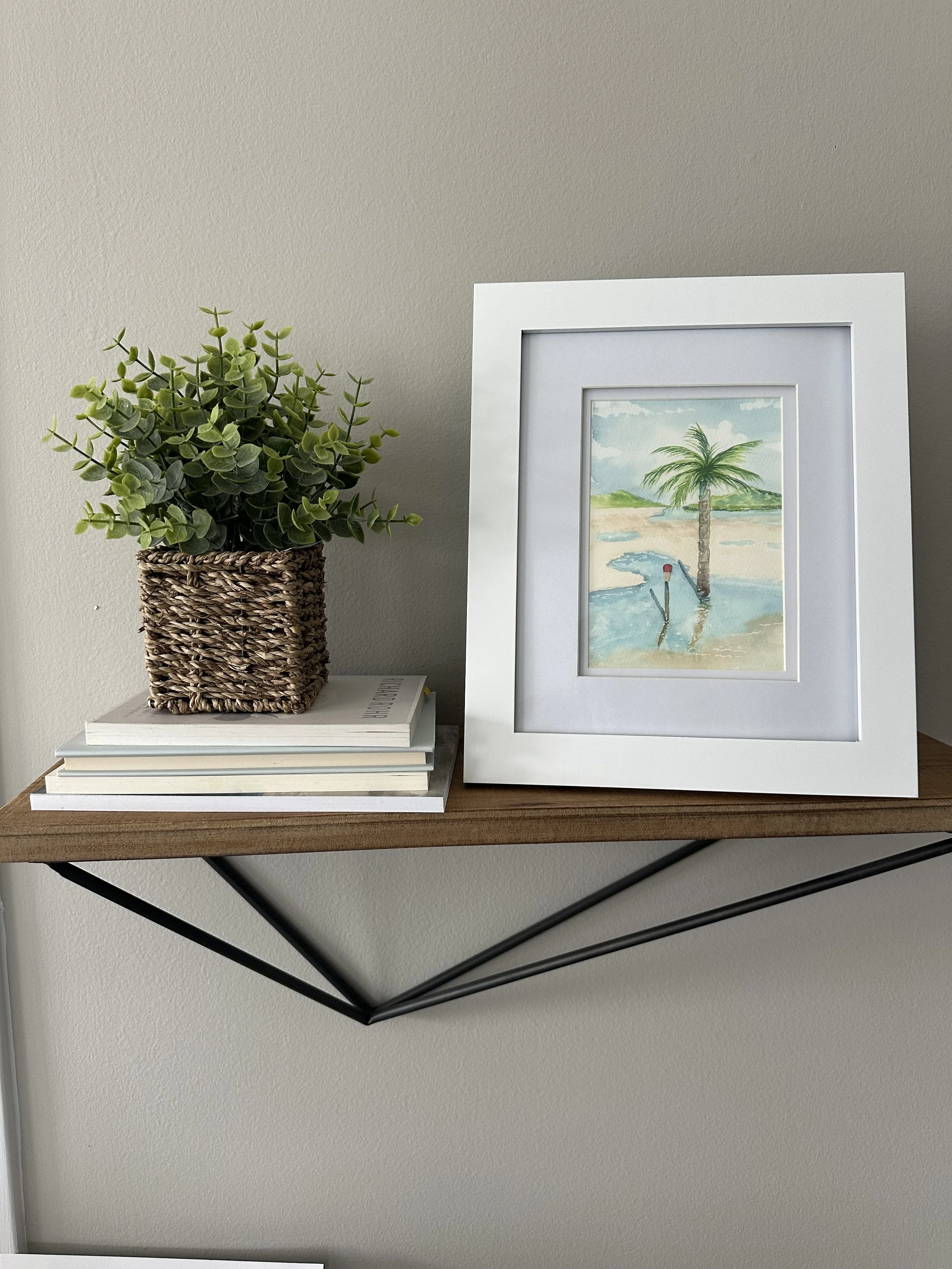 A wooden shelf with black metal supports holds a potted green plant, a stack of four books, and a framed watercolor painting of a beach scene with a palm tree and water.