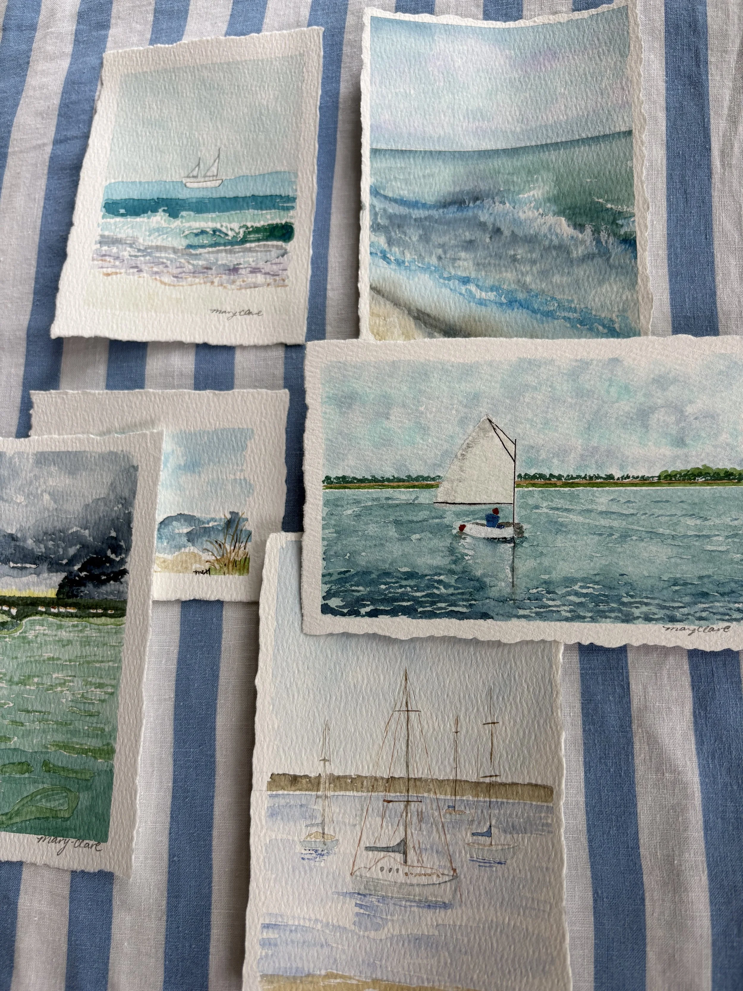 Collection of watercolor paintings of maritime scenes including sailboats, ocean waves, stormy skies, and coastal landscapes laid out on a striped fabric background.