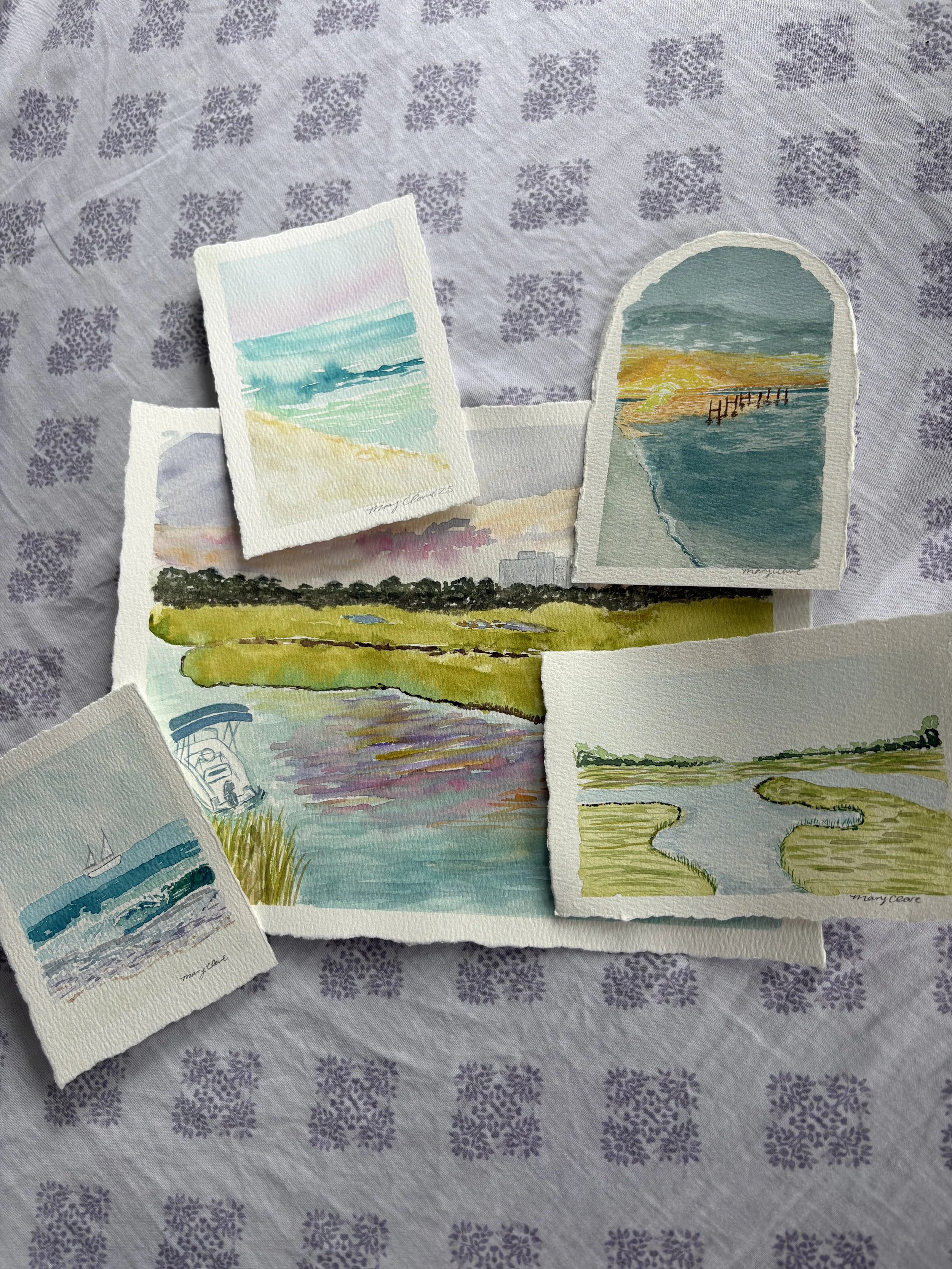 Collection of small watercolor paintings of coastal and marsh scenes, arranged on a patterned tablecloth.