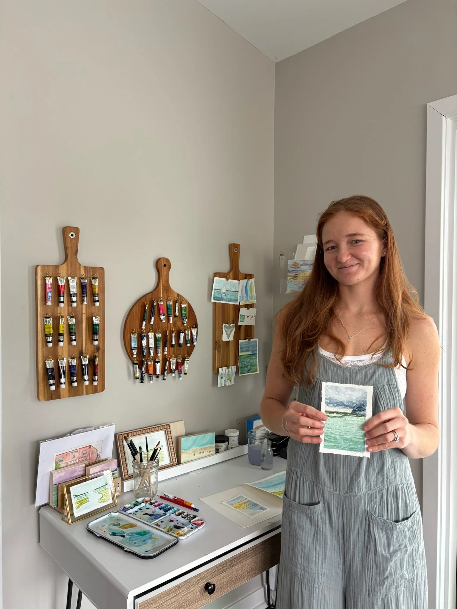 A woman with long red hair standing in an art studio, holding a small landscape painting. The studio has watercolors, paintbrushes, and small framed artworks on a white desk with wall-mounted wooden display racks with tubes of paint.