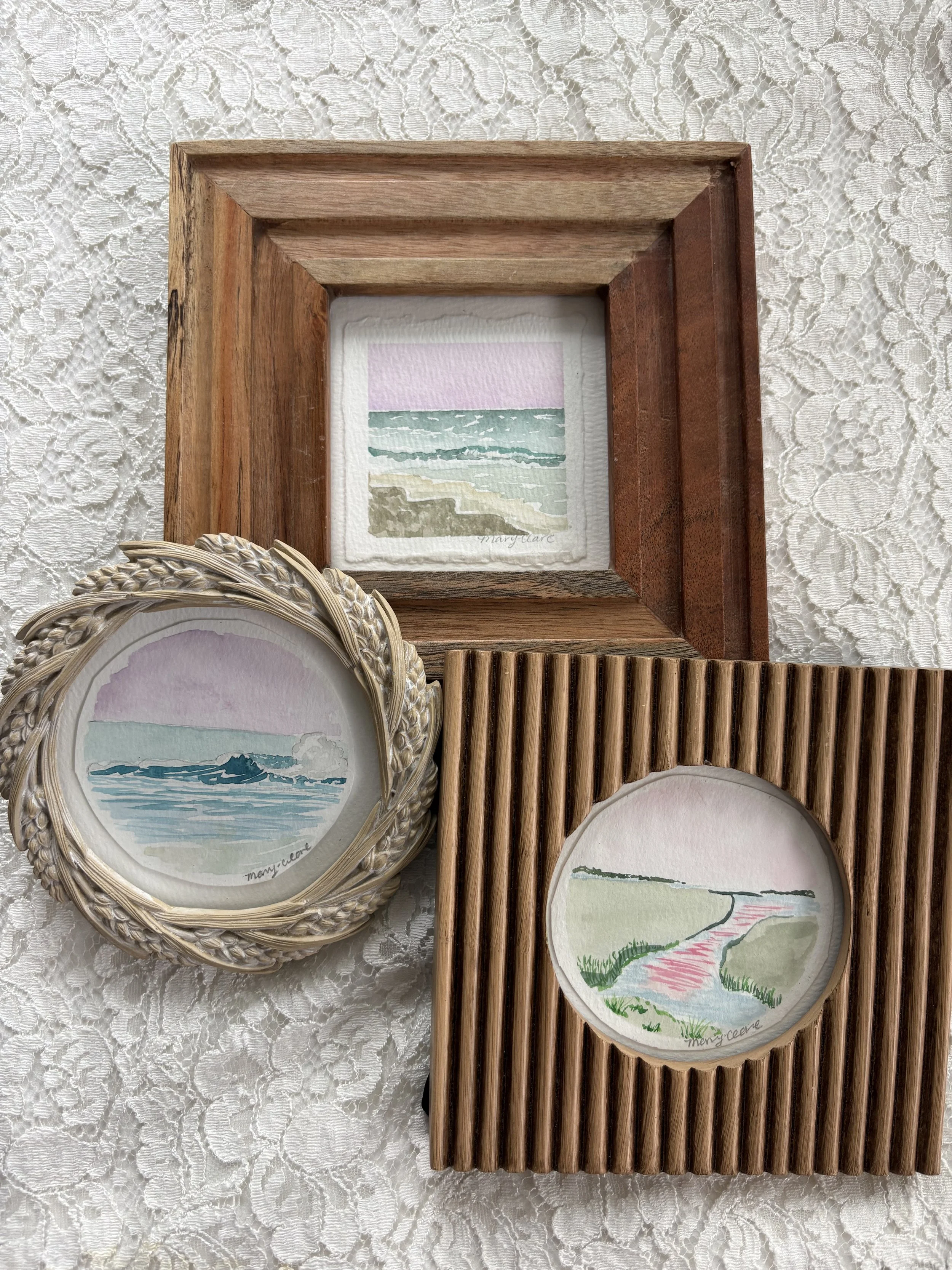 Four watercolor paintings of beach scenes are displayed on a lace tablecloth, each framed differently.