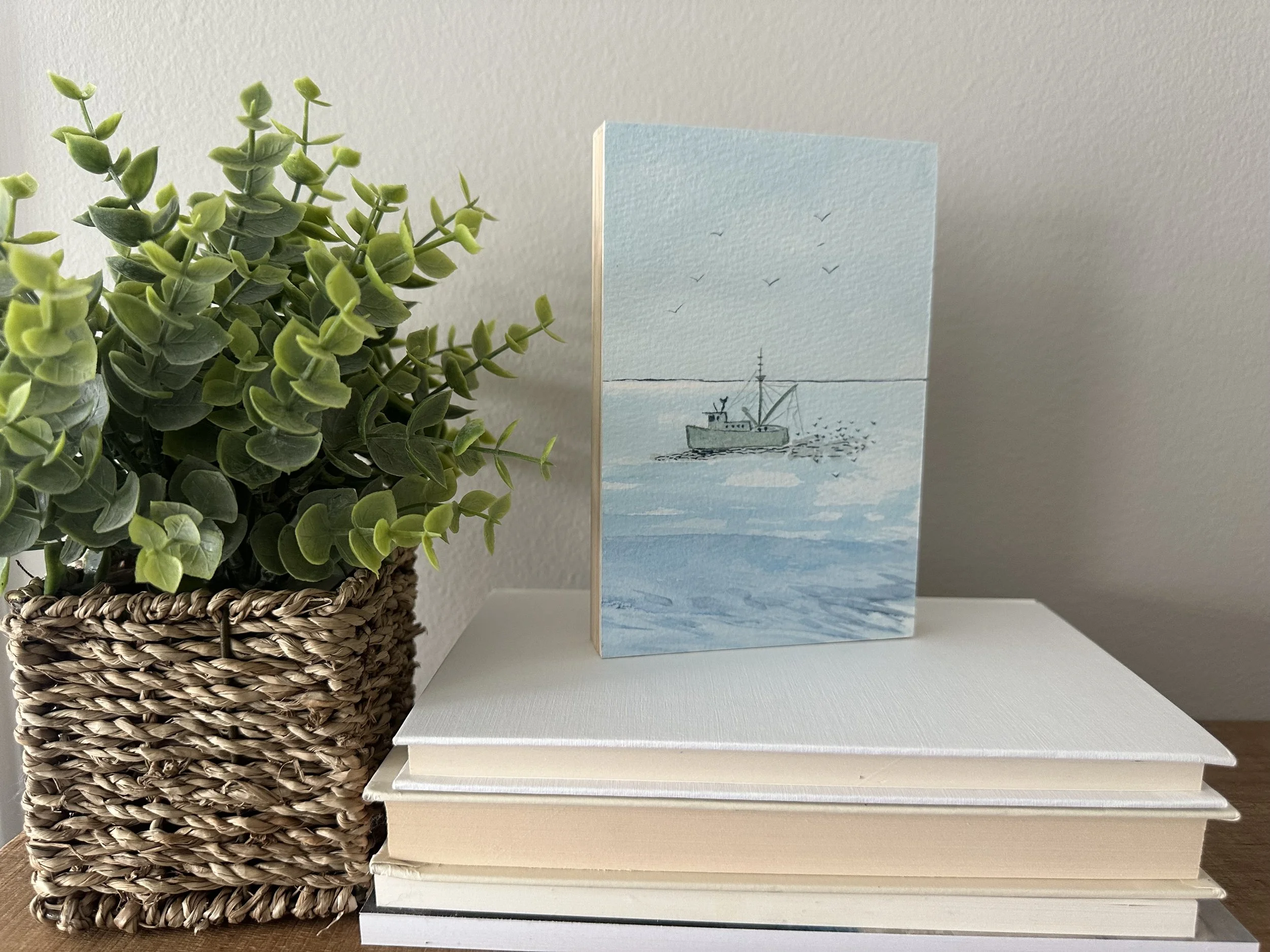 A potted green plant with leafy foliage on a wooden surface next to a stack of books and a card with a blue watercolor scene of a boat on water and birds flying above.