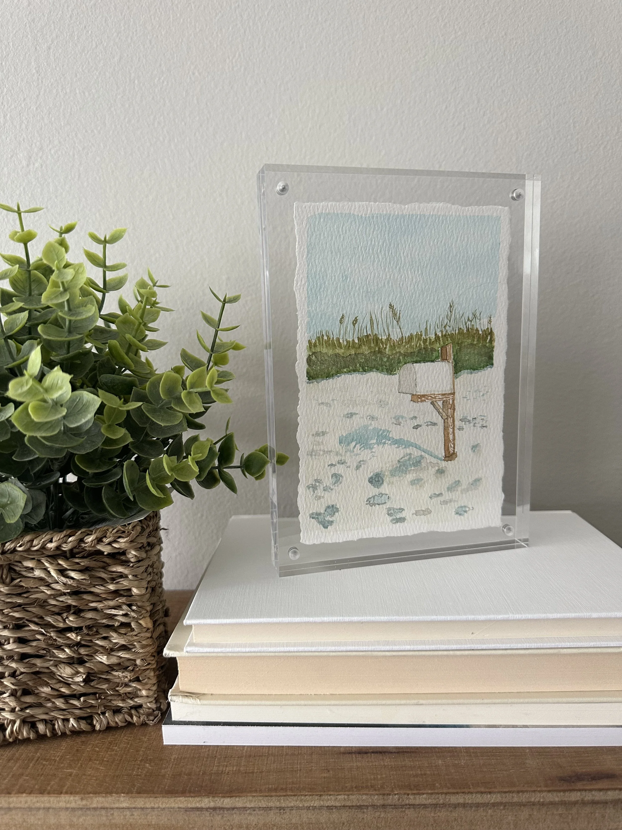 Watercolor painting of a snow-covered field with a mailbox and a line of tall grass or reeds in the background, enclosed in a clear acrylic frame, placed on books next to a potted green plant.