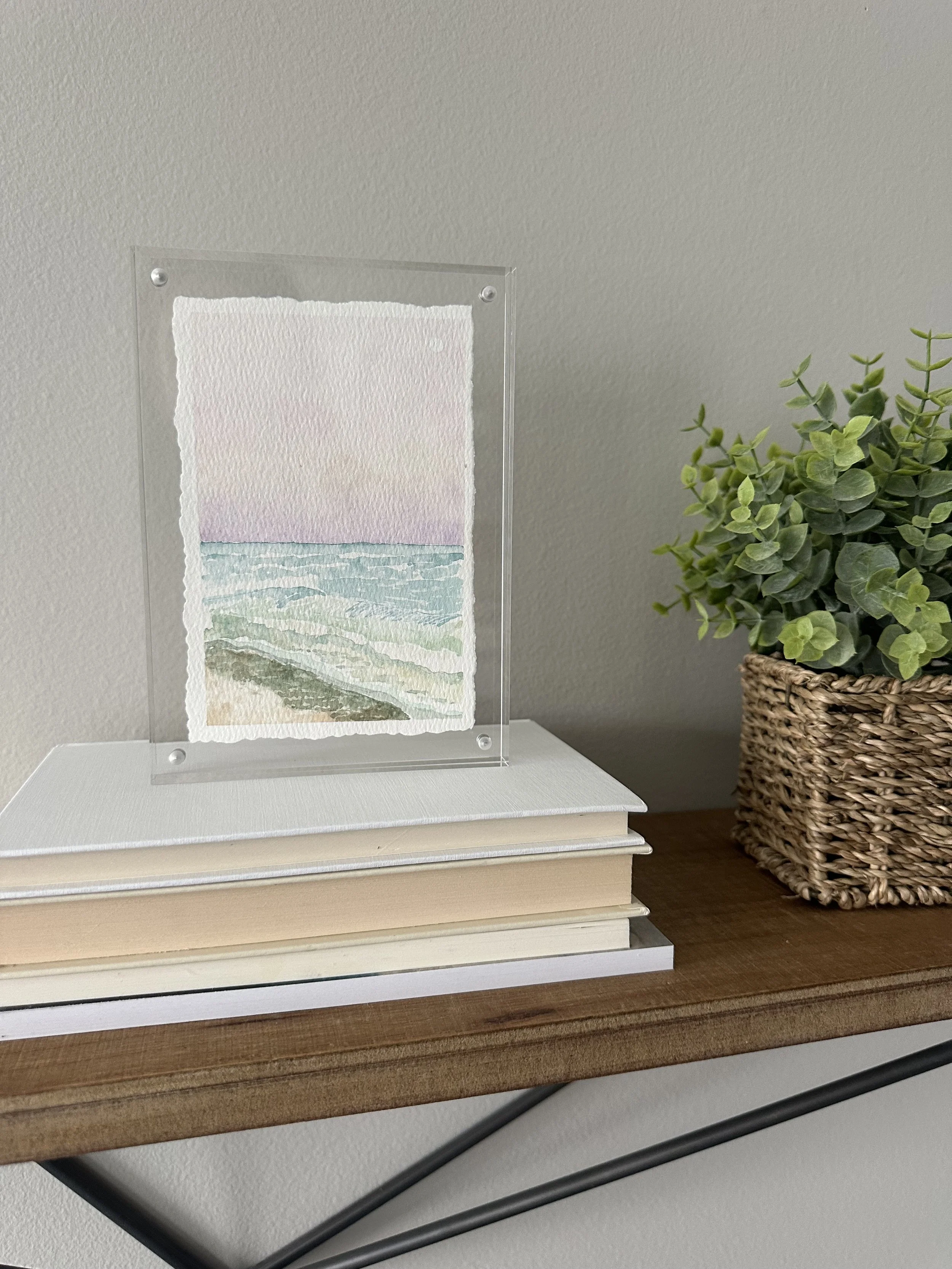 A watercolor painting of the ocean, with gentle waves and a pink sky, resting on top of a stack of books on a wooden shelf.