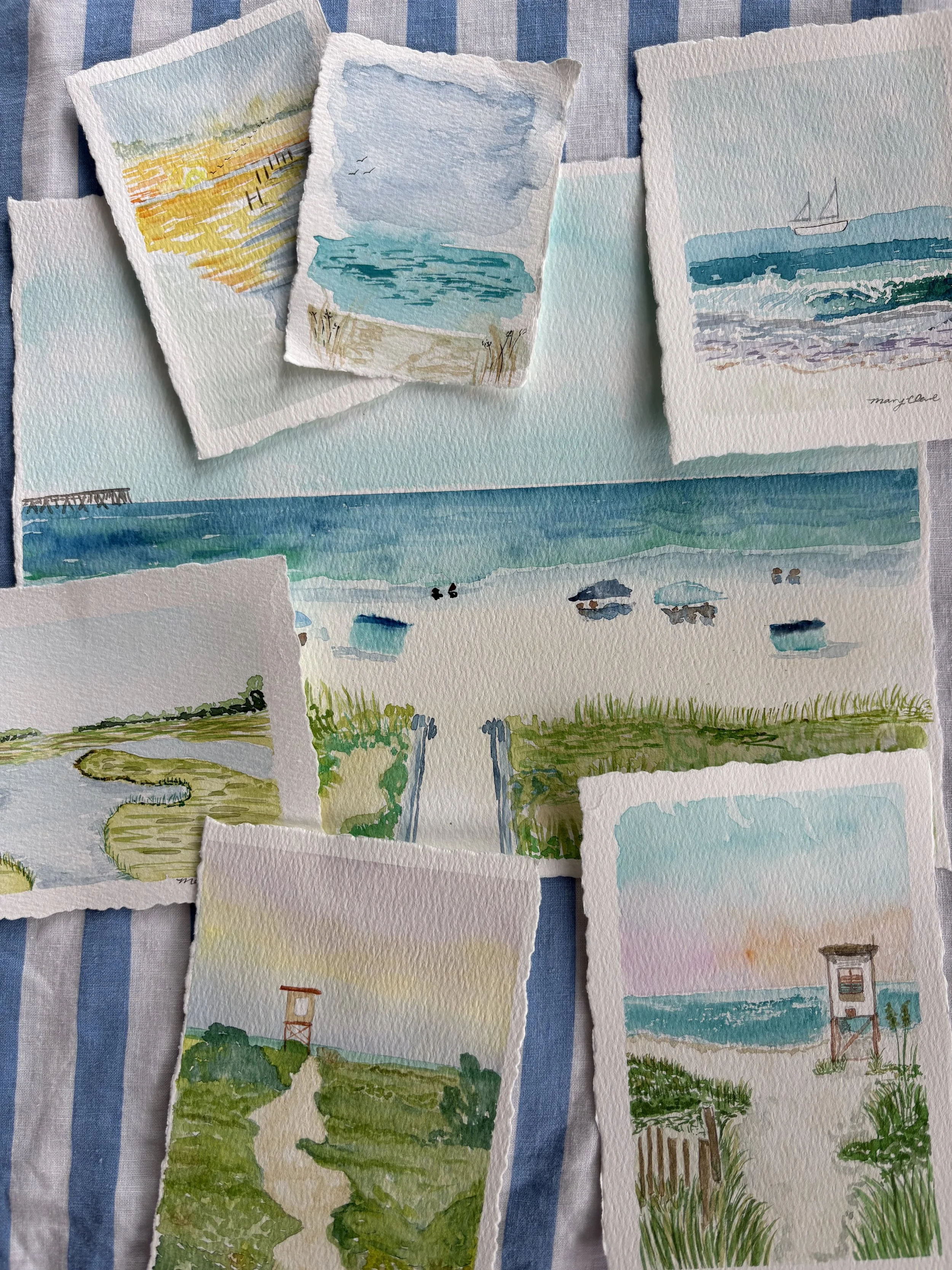 Collection of watercolor paintings of beach scenes, including ocean waves, boats, a pier, grassy paths, and a lifeguard tower, arranged on a blue-and-white striped fabric background.