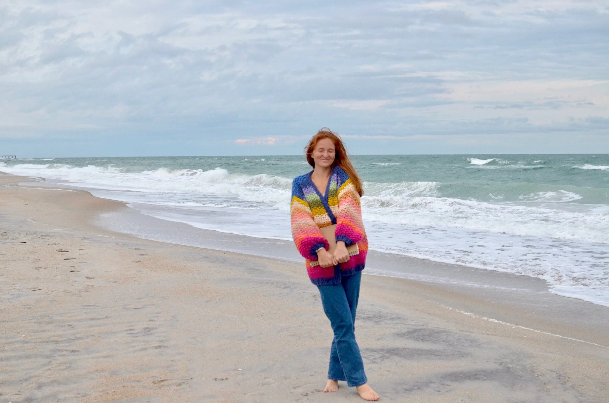 A young woman with red hair is smiling and walking barefoot on a sandy beach near the ocean, wearing a colorful striped sweater and blue jeans.