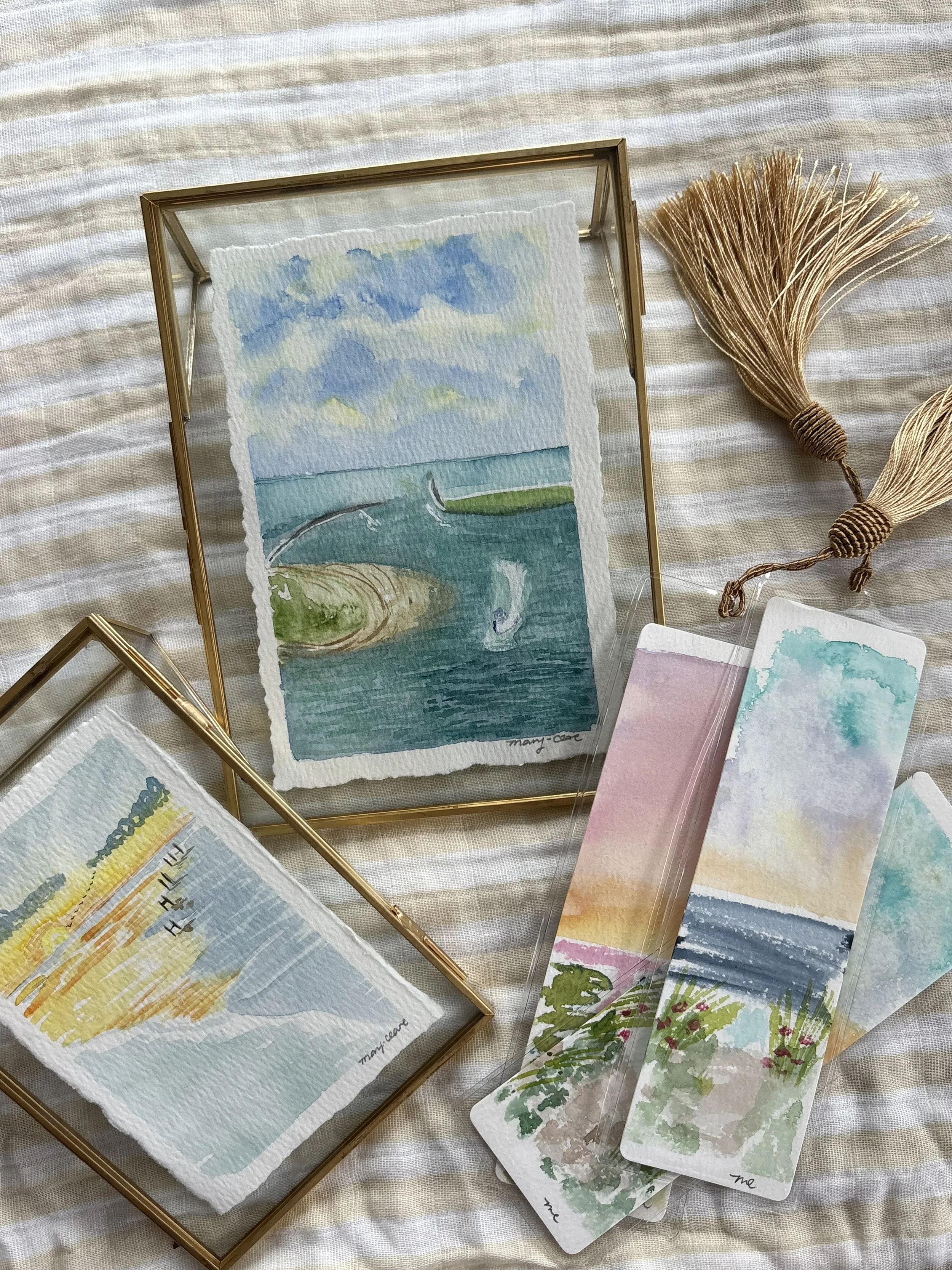 Collection of watercolor paintings of landscapes and seascapes, framed in glass and gold, with decorative tassels on a striped cloth.
