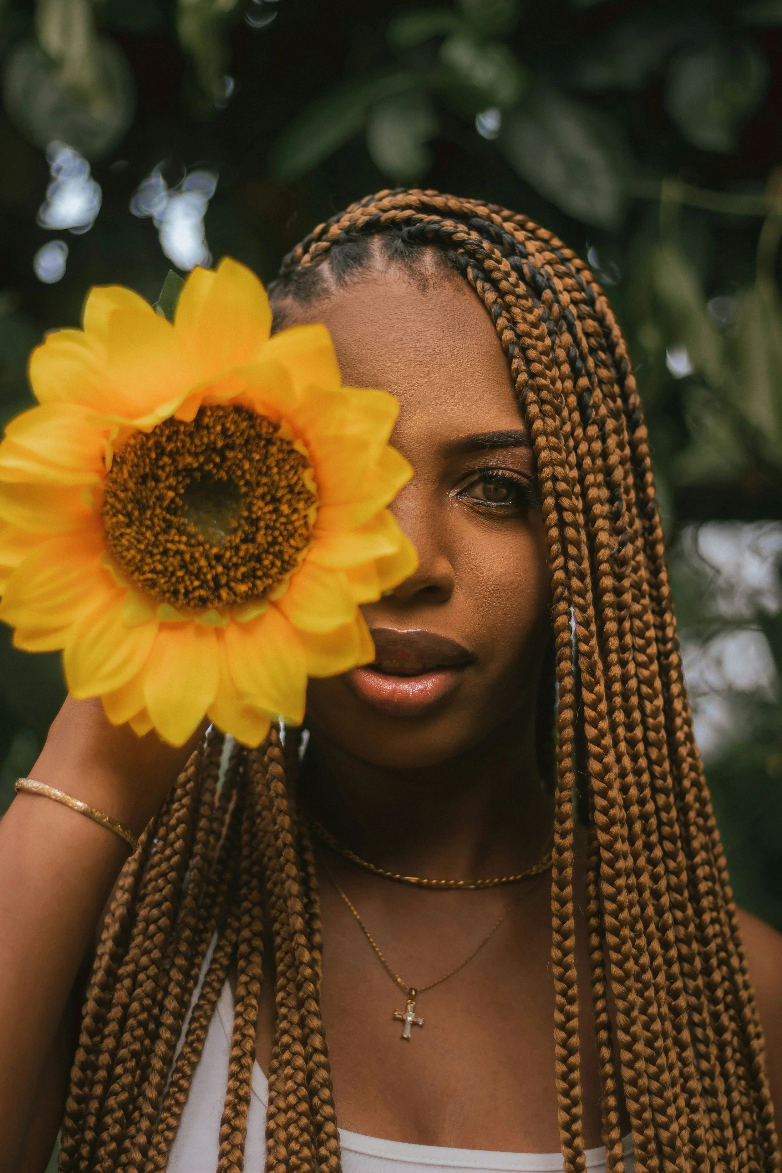 A woman with long, braided hair holding a sunflower partially covering her face outdoors.