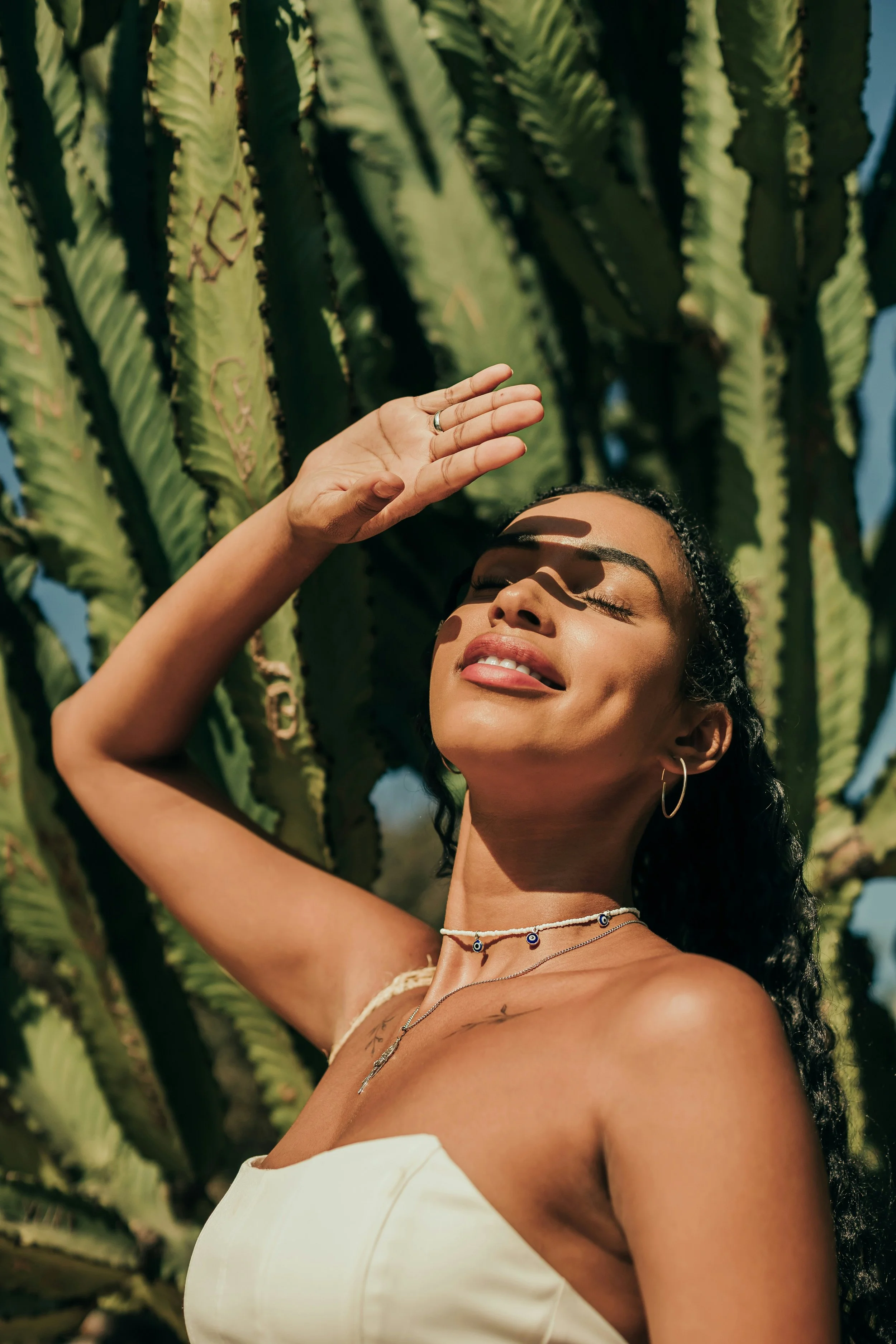 A woman with curly hair and hoop earrings tilts her face up with her eyes closed, shielding her face from the sun with her hand, standing in front of a cactus in bright sunlight.