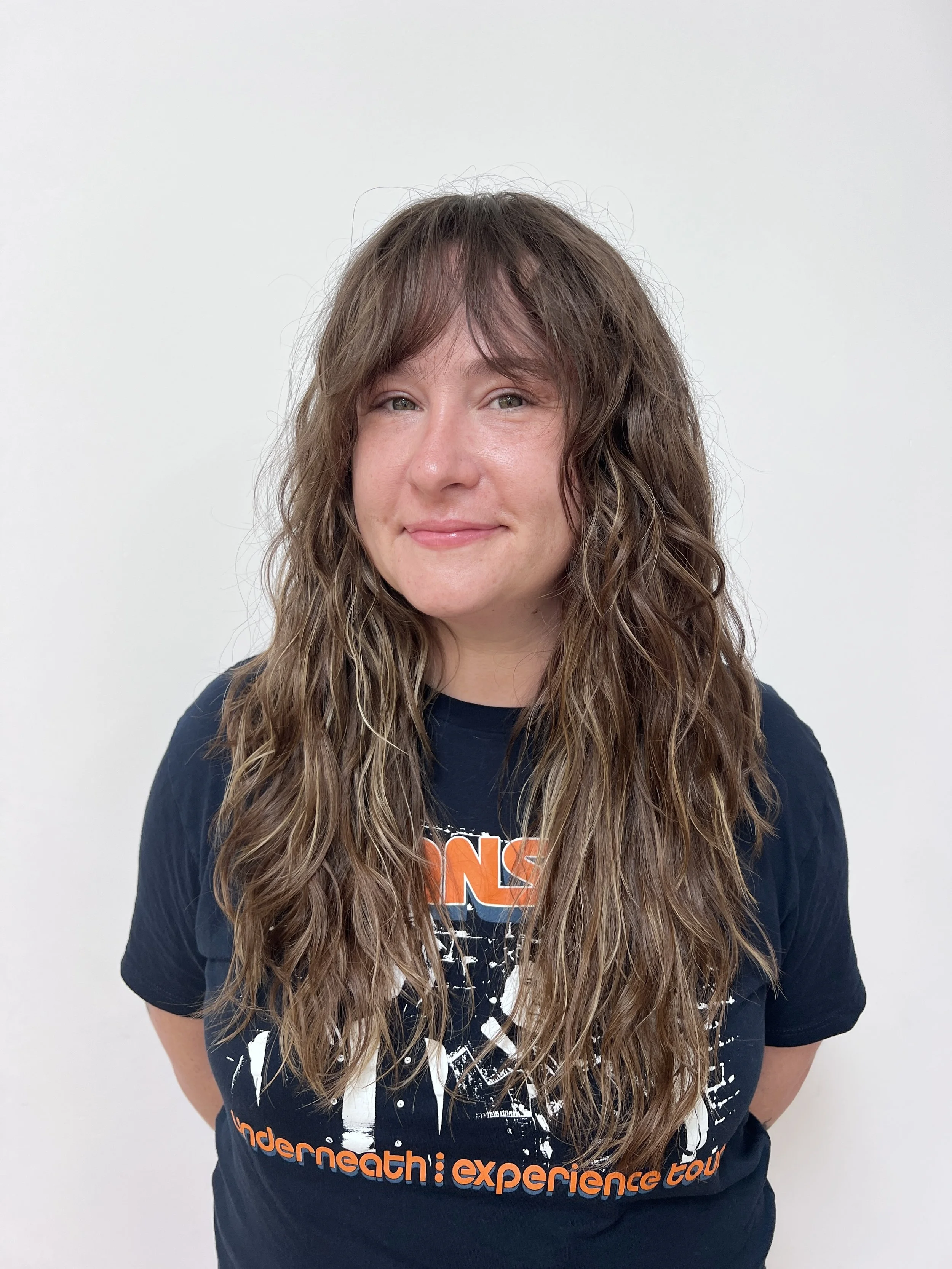 A woman with long, wavy brown hair and light skin, smiling slightly, wearing a black T-shirt with orange and white graphics and text, standing against a plain white background.