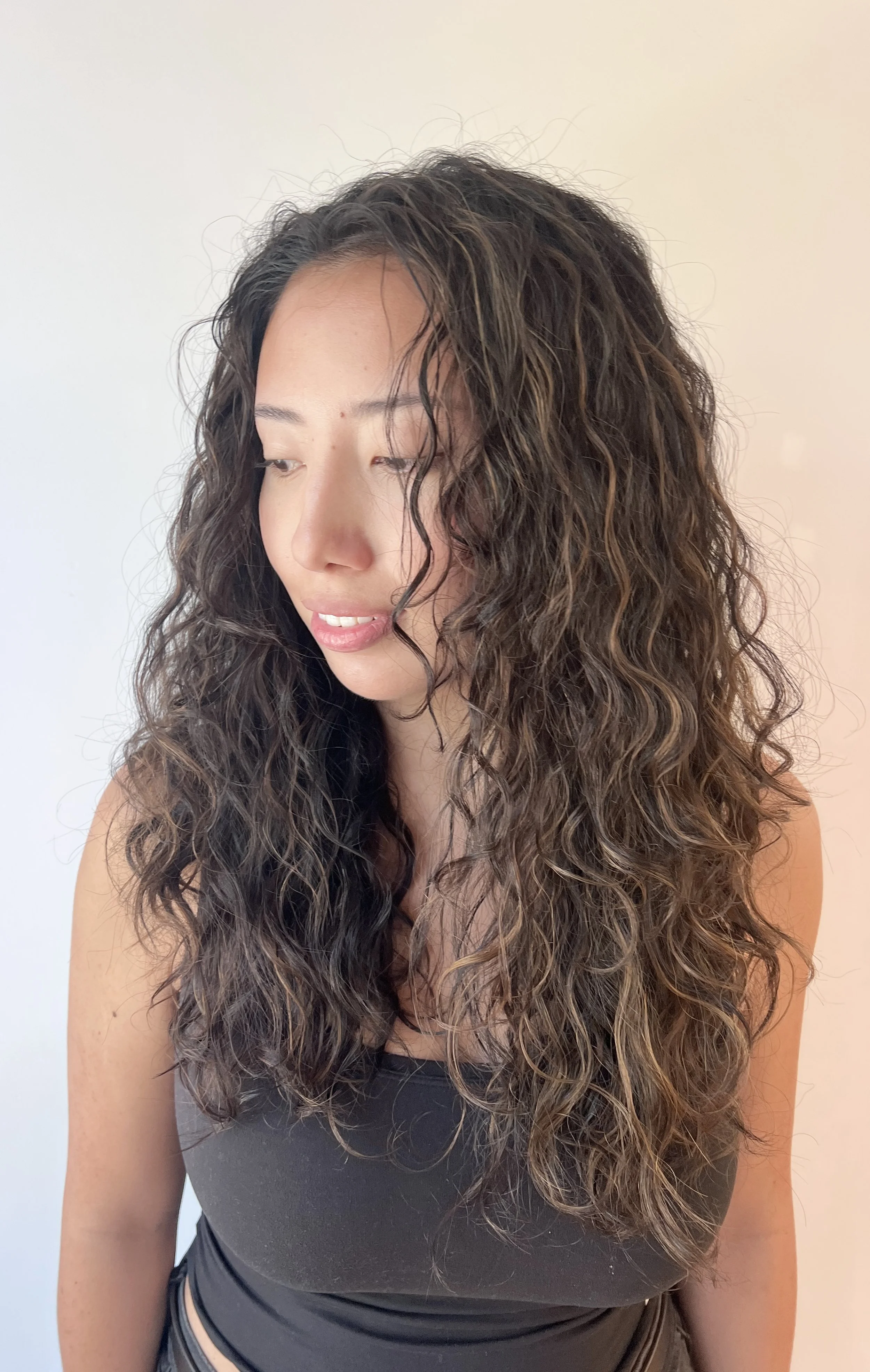 A young woman with long, curly hair wearing a black crop top, looking down and to the side against a plain white background.