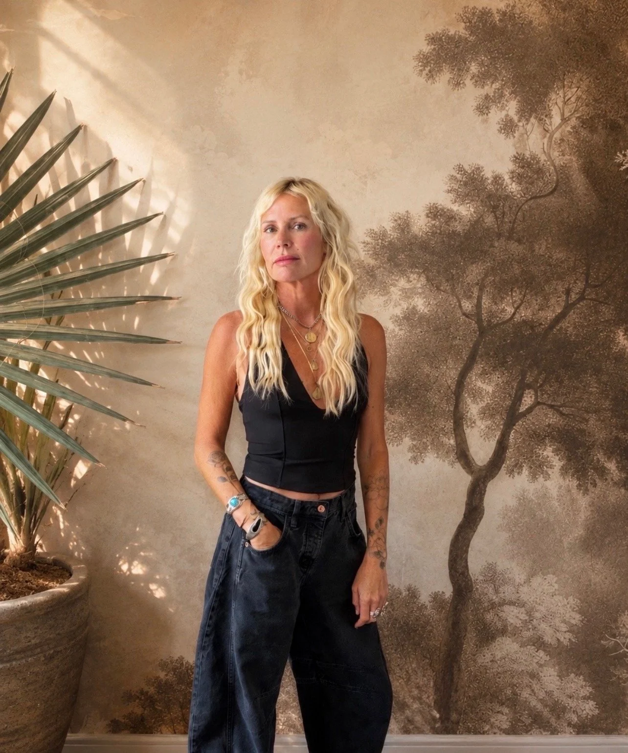 blonde hair stylist with gold jewelry and black vest, black jeans stands with hand in pocket. Plants and greenery are around her.