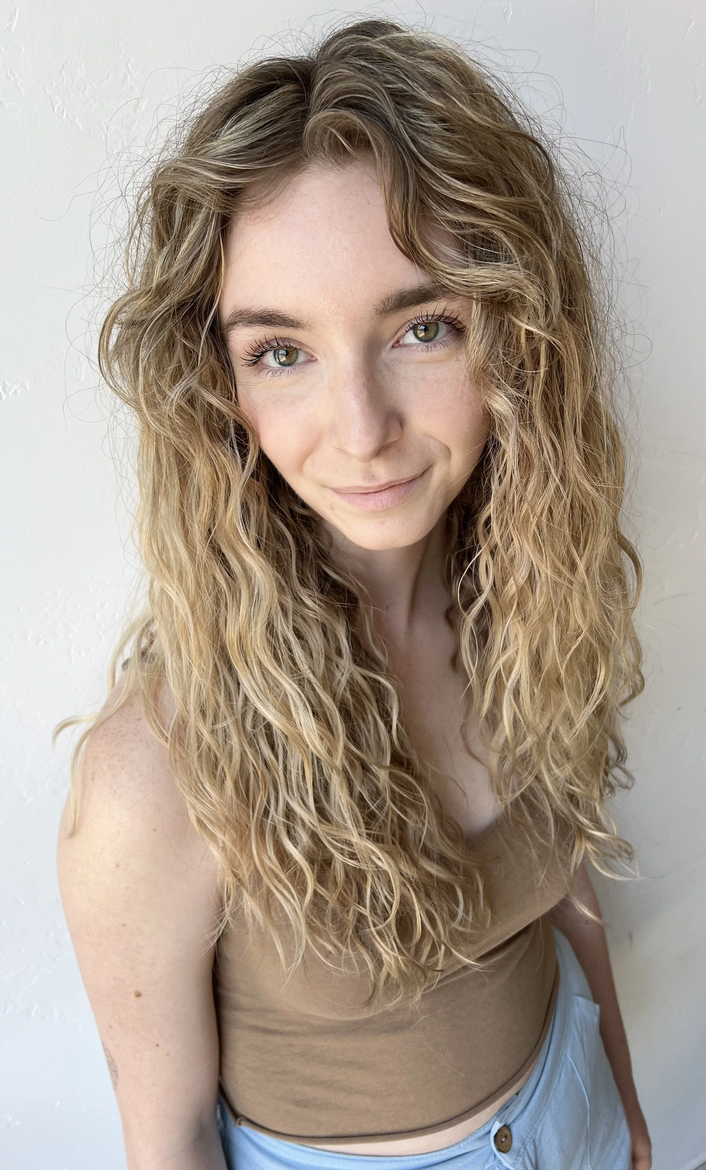 A young woman with long, curly blonde hair and light-colored eyes, smiling softly while looking directly at the camera. She is wearing a tan sleeveless top and light blue shorts, standing against a plain, light-colored wall.