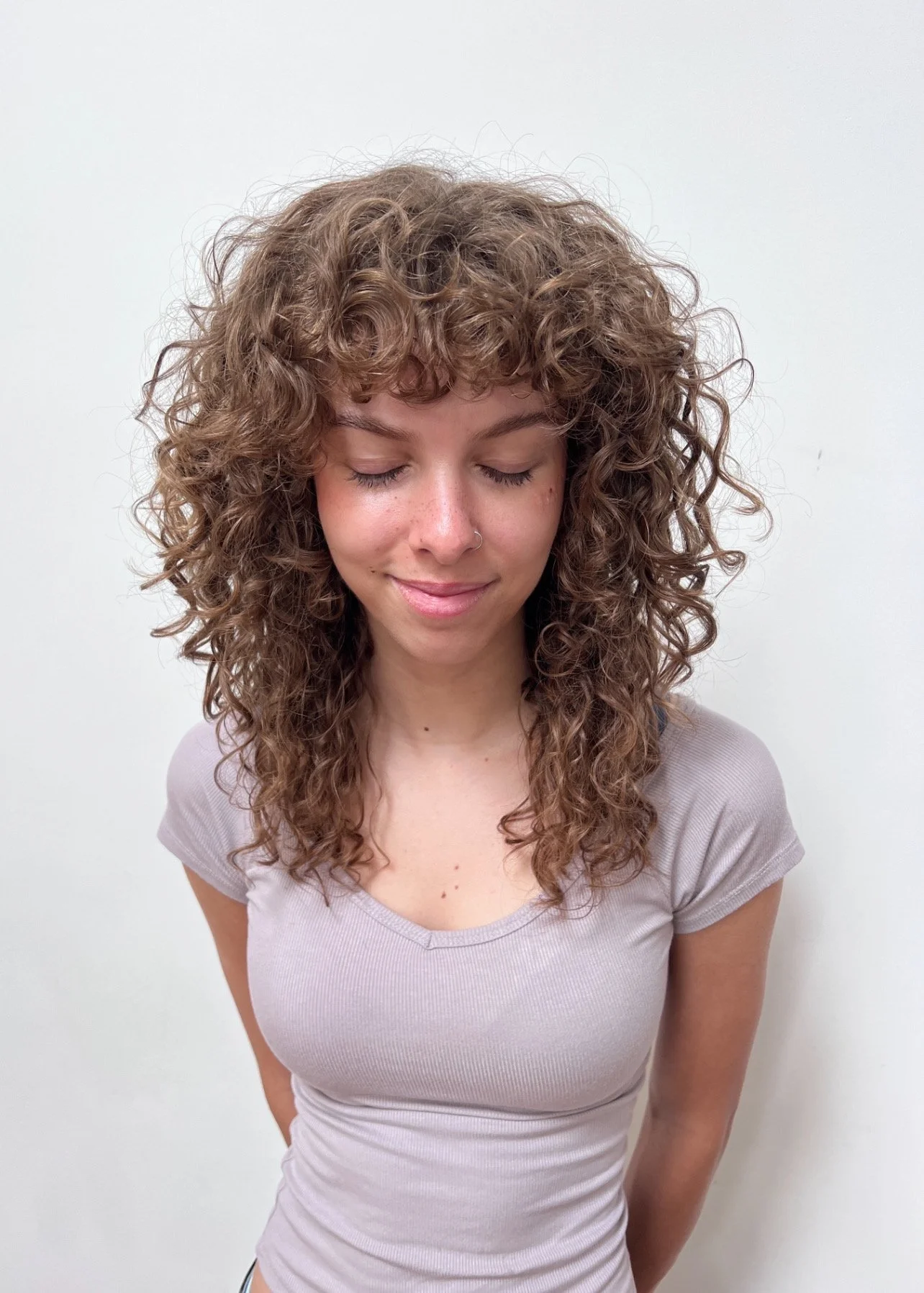 A woman with curly brown hair, wearing a light purple t-shirt, has her eyes closed and a slight smile, standing against a plain white wall.